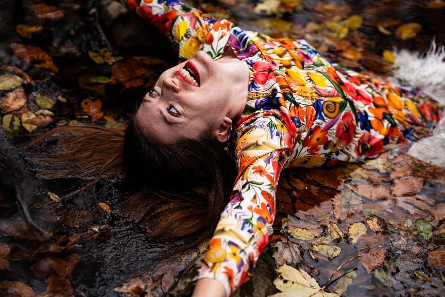 Someone in a colorful floral dress lies on autumn leaves with long hair spread out on the ground.