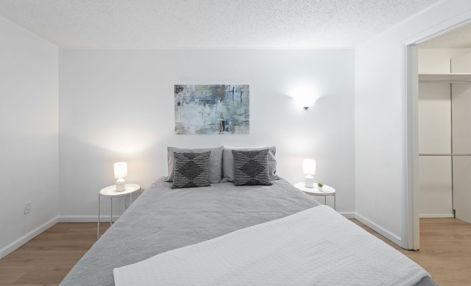 Modern white bedroom with gray bedding, decorative pillows, matching nightstands, wall lamps, and hardwood floors.