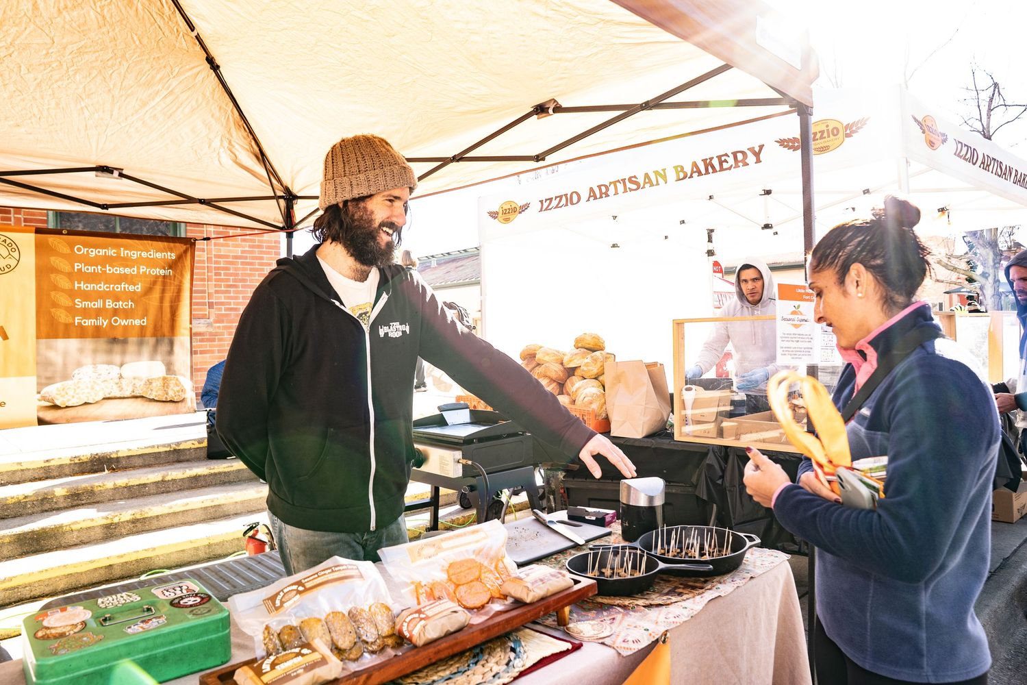 Man laughing while talking to a customer about samples at The Little House of Tempeh booth at the farmers market.
