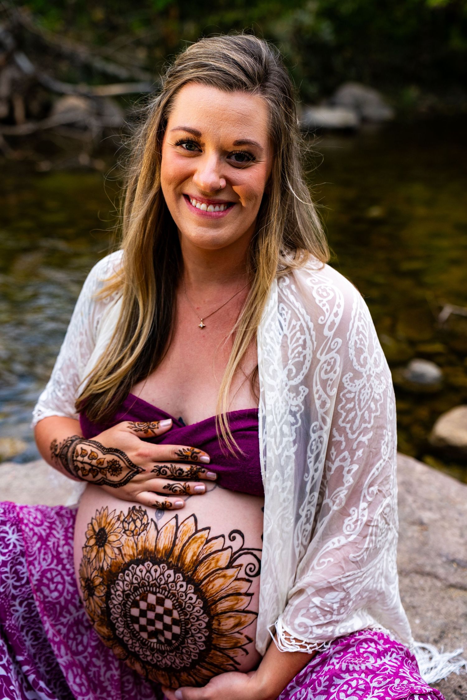 Woman in purple dress and white lace cardigan sits by river rocks during outdoor photo session.