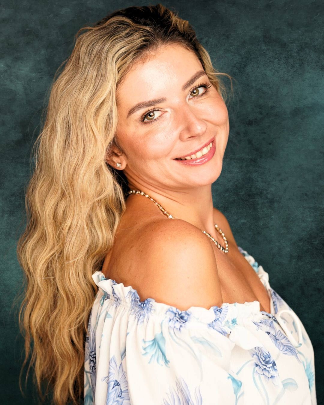 Smiling blonde woman in floral off-shoulder top with wavy hair against teal backdrop.