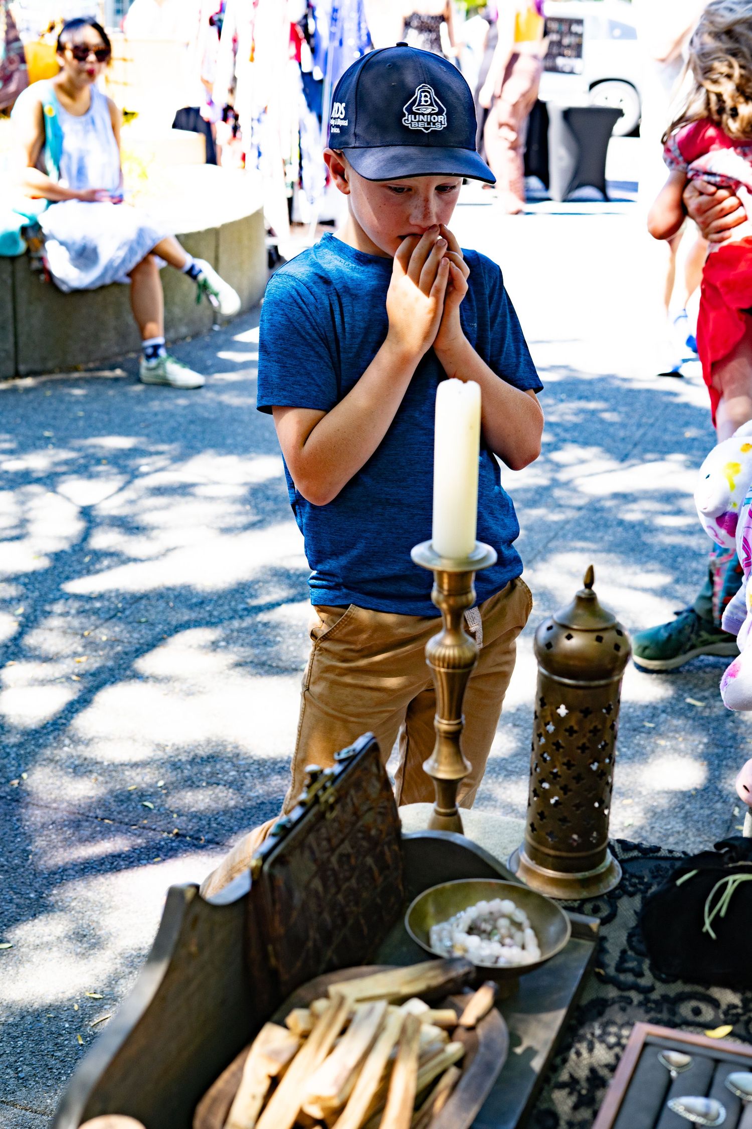Boy in blue shirt and cap examines brass candlestick and lantern at outdoor market display.