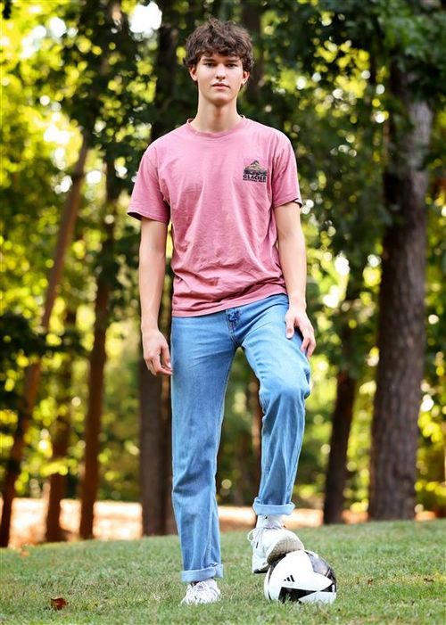 Teen boy in pink t-shirt and jeans standing outdoors with soccer ball at feet near trees.
