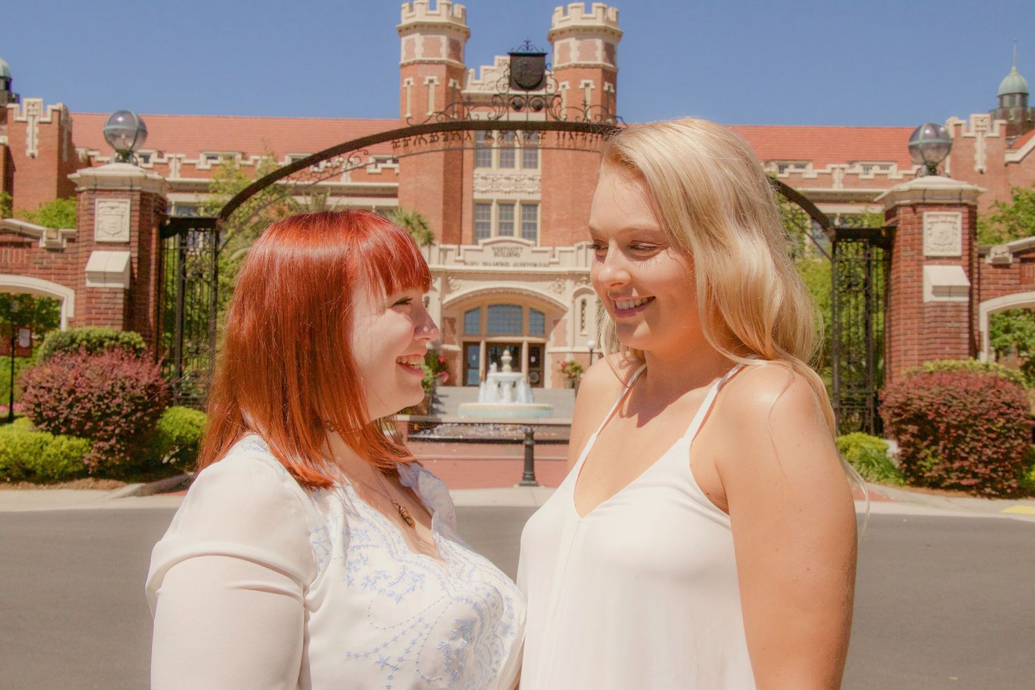 Two young women, one redhead and one blonde, smiling at each other in front of a red brick university building.