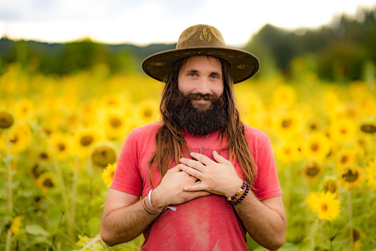 Bearded man with long hair and wide-brim hat smiling in a vibrant sunflower field, wearing a red t-shirt.