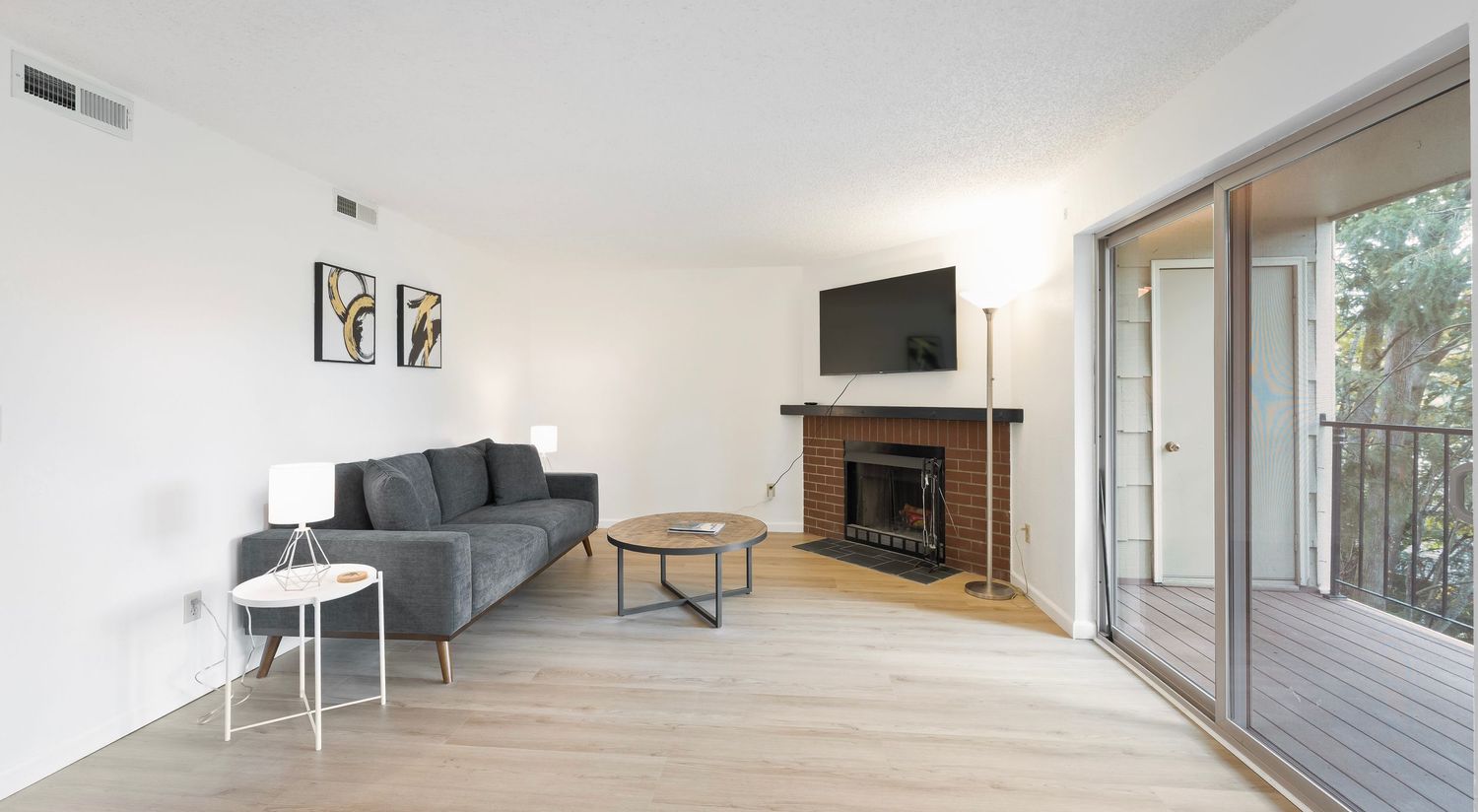 Modern living room with gray sofa, brick fireplace, wall-mounted TV, and sliding glass doors to a balcony.
