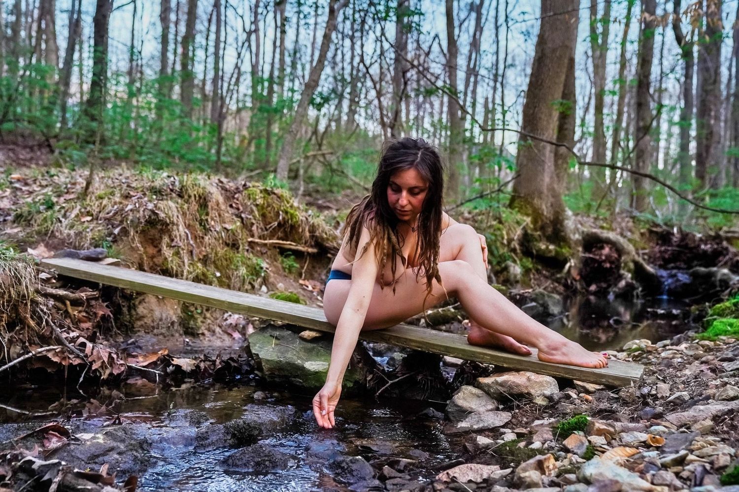 A photo of a woman half naked touching the water in the river. There are lots of trees behind her with many stones and other nature.