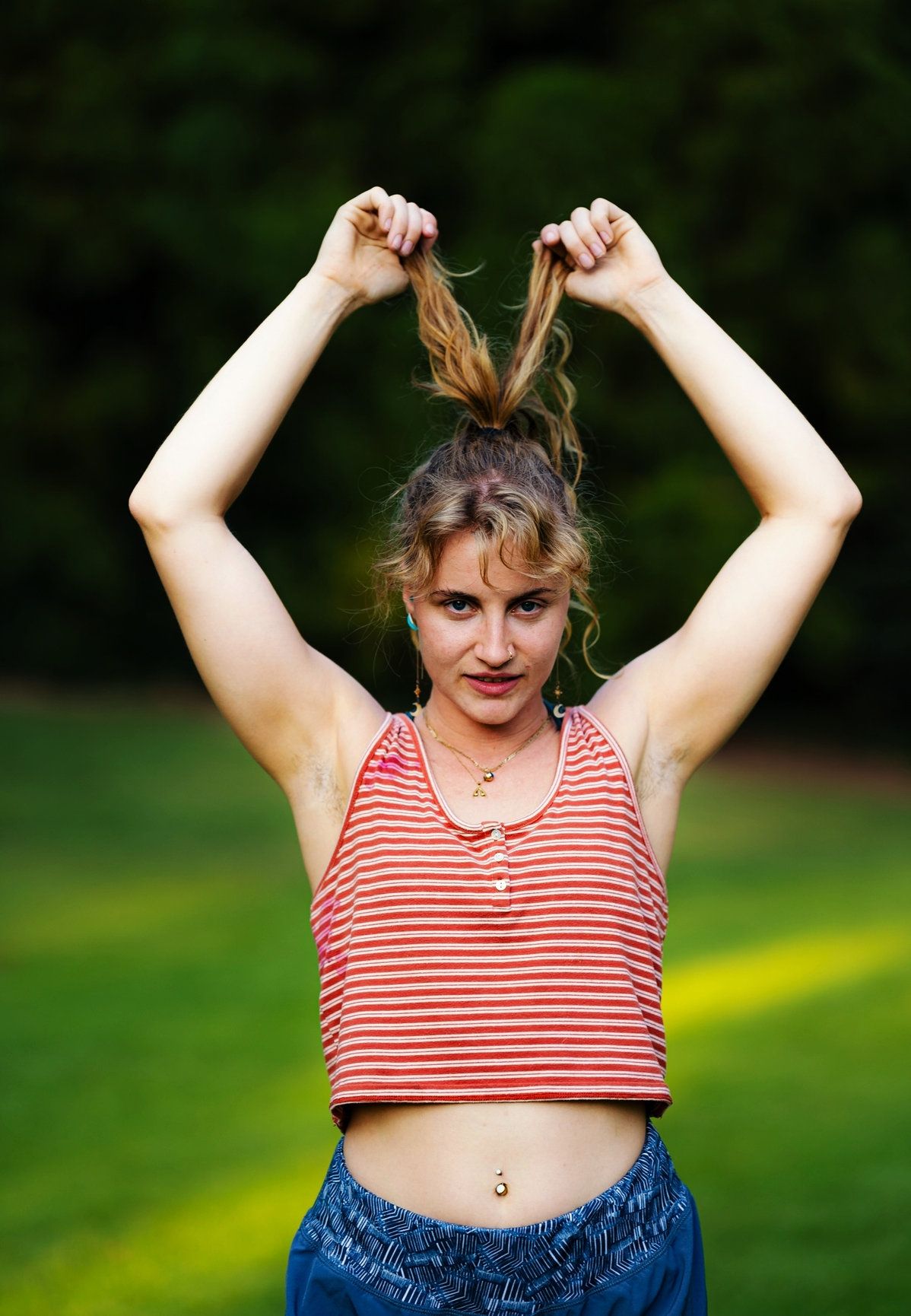 A photo of a blonde woman fixing her hair wearing a red striped tank top with blue pants.