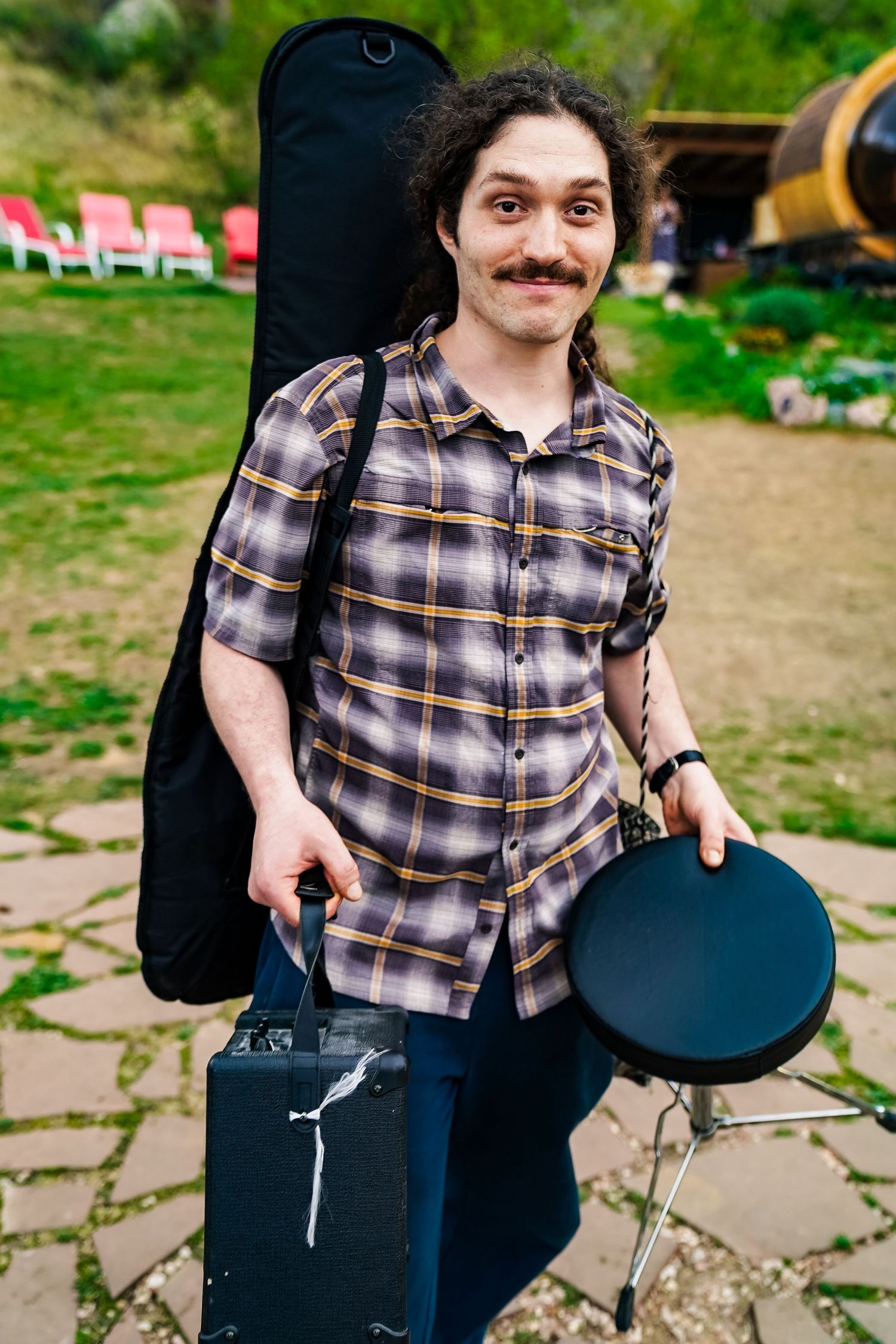 Young musician with curly hair and mustache carrying guitar case, amp, and drum pad outdoors.
