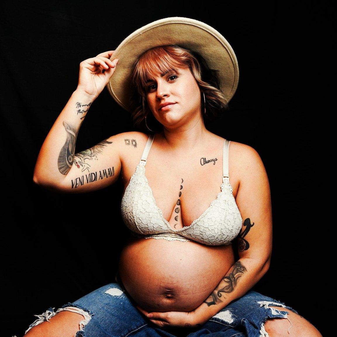 A pregnant woman in a studio with a black backdrop, holding her belly and her hat. The photo conveys elegance, anticipation, and a serene.