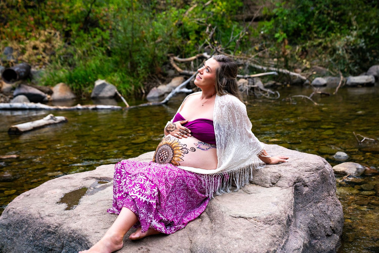 A person in a purple dress relaxes on large rocks by a flowing river on a sunny summer day.