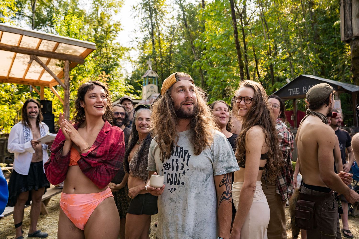 Group of festival-goers enjoying an outdoor music festival in a wooded area on a sunny day.