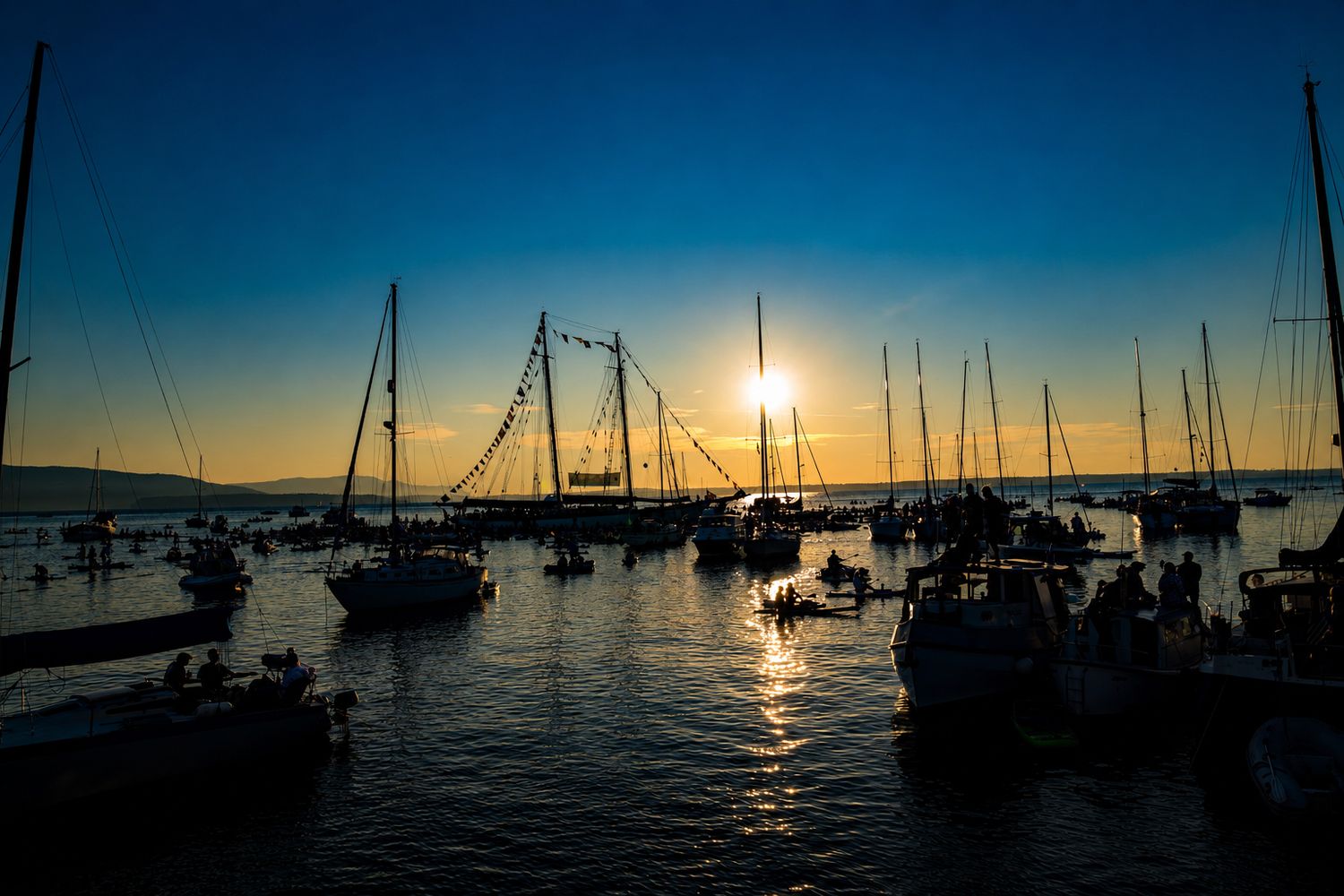 Silhouetted sailboats and yachts in a marina at golden sunset with vibrant blue sky reflecting on calm water.