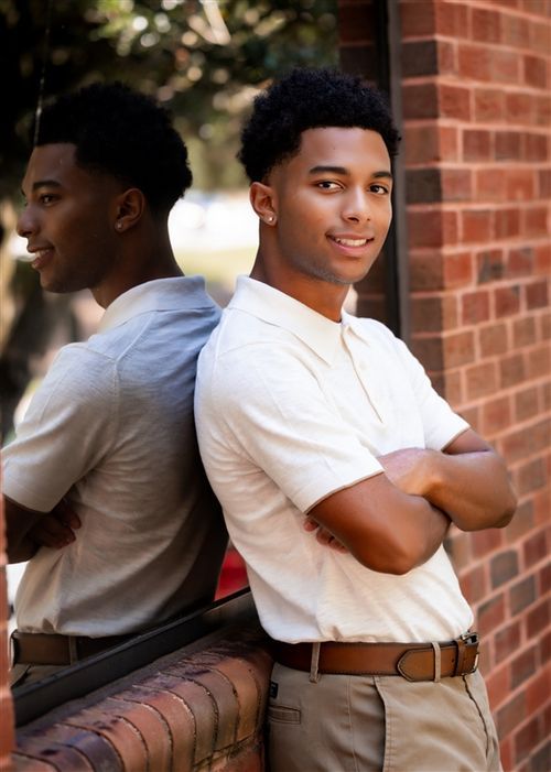 Two young men in casual attire lean back-to-back against a brick wall, smiling confidently outdoors.