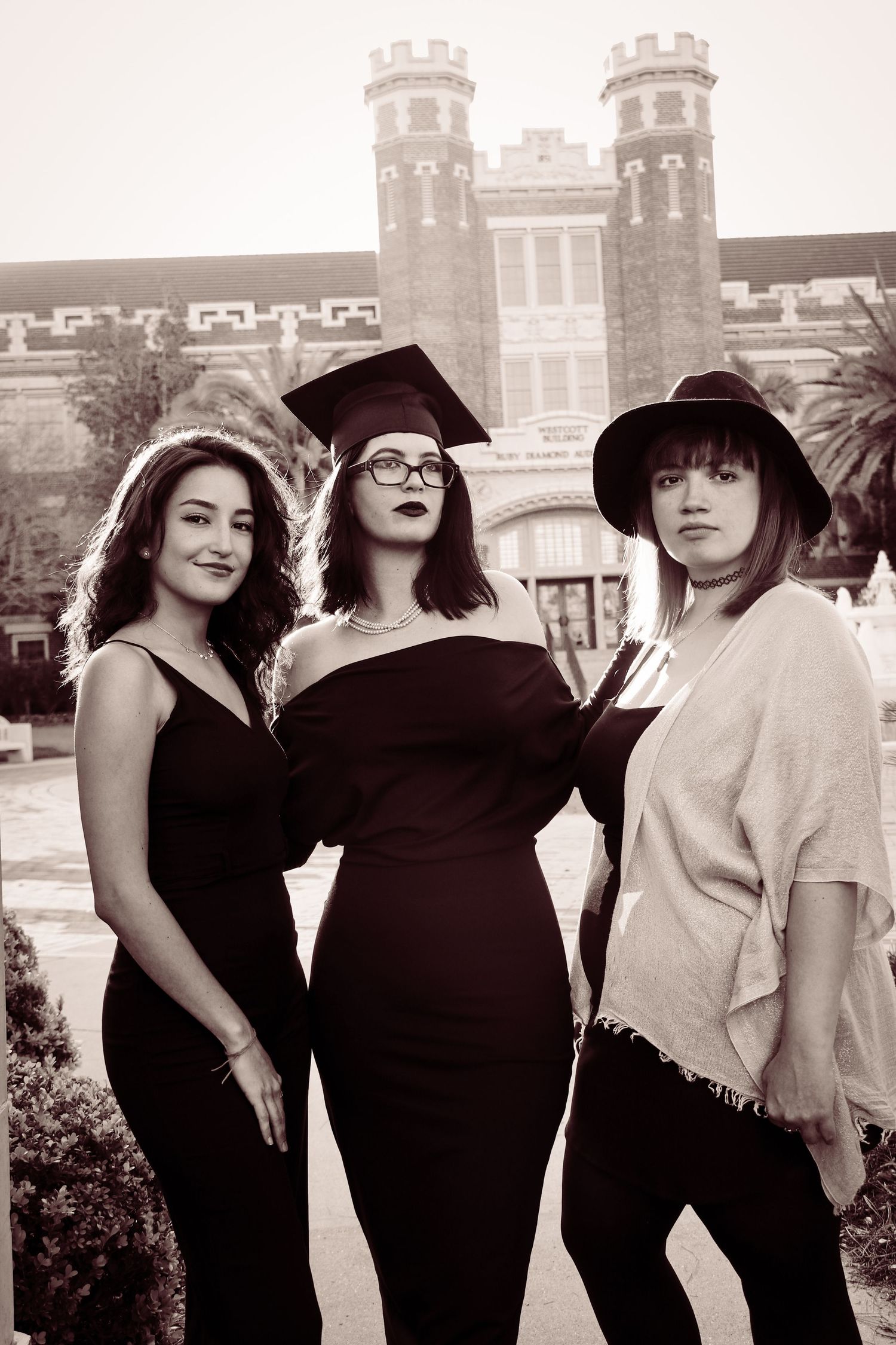Three young women pose outdoors near a brick building, one wearing a graduation cap and gown in a sepia-toned photo.