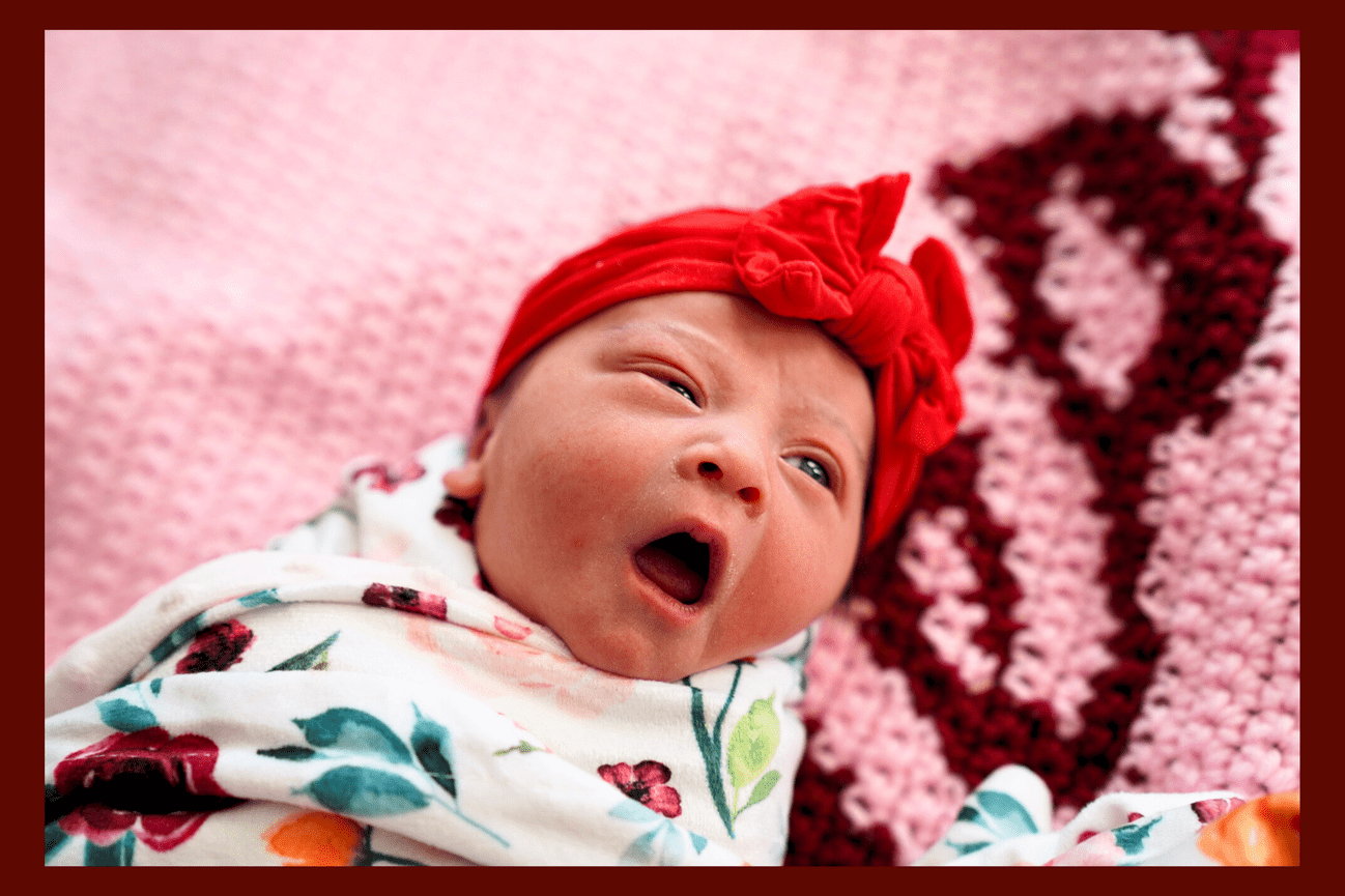 A photo of a baby yawning, wrapped up in a blanket wearing a red bowtie on her head.