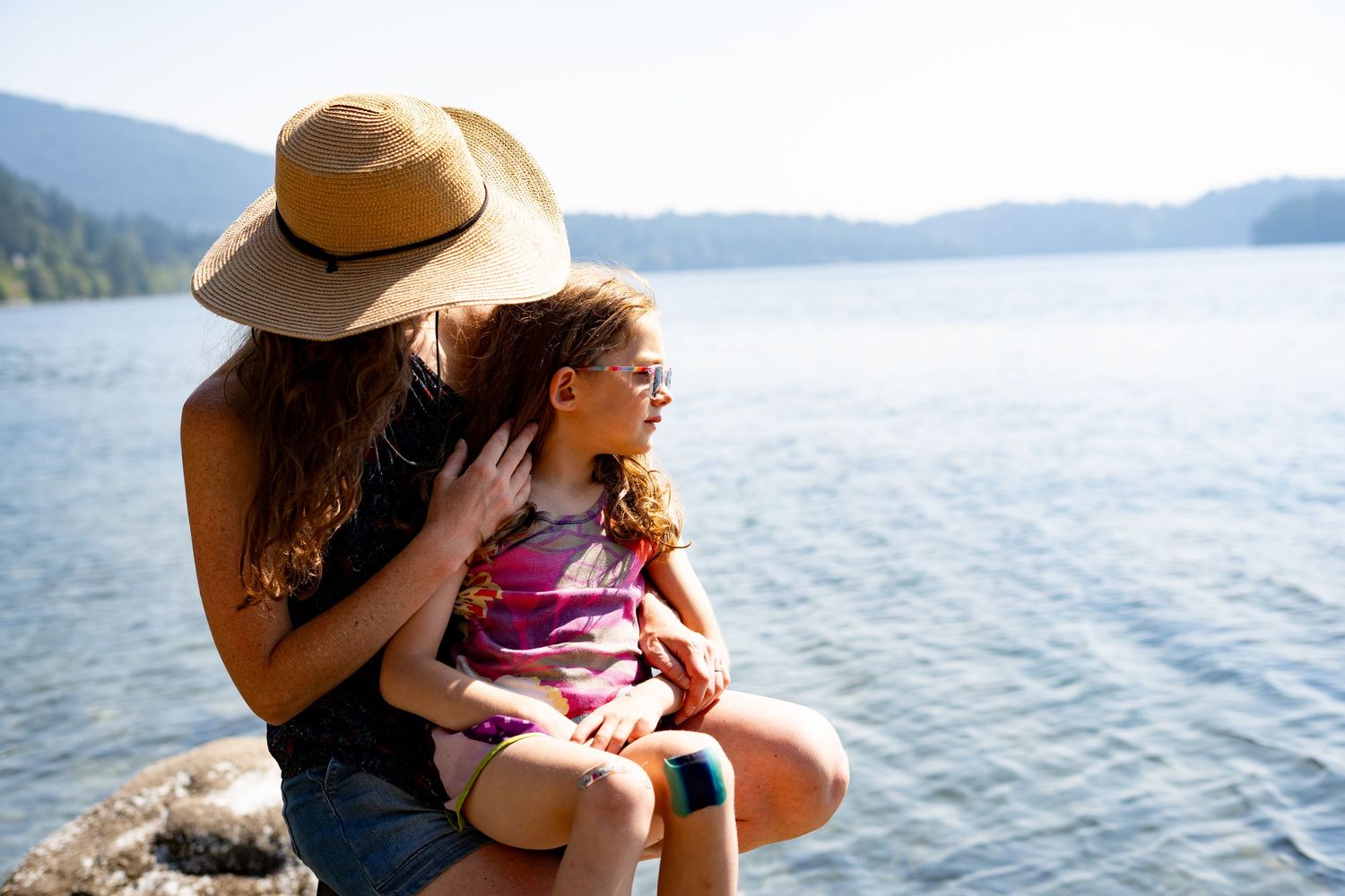 A mother and young daughter sit together on rocks by a serene mountain lake on a sunny day.