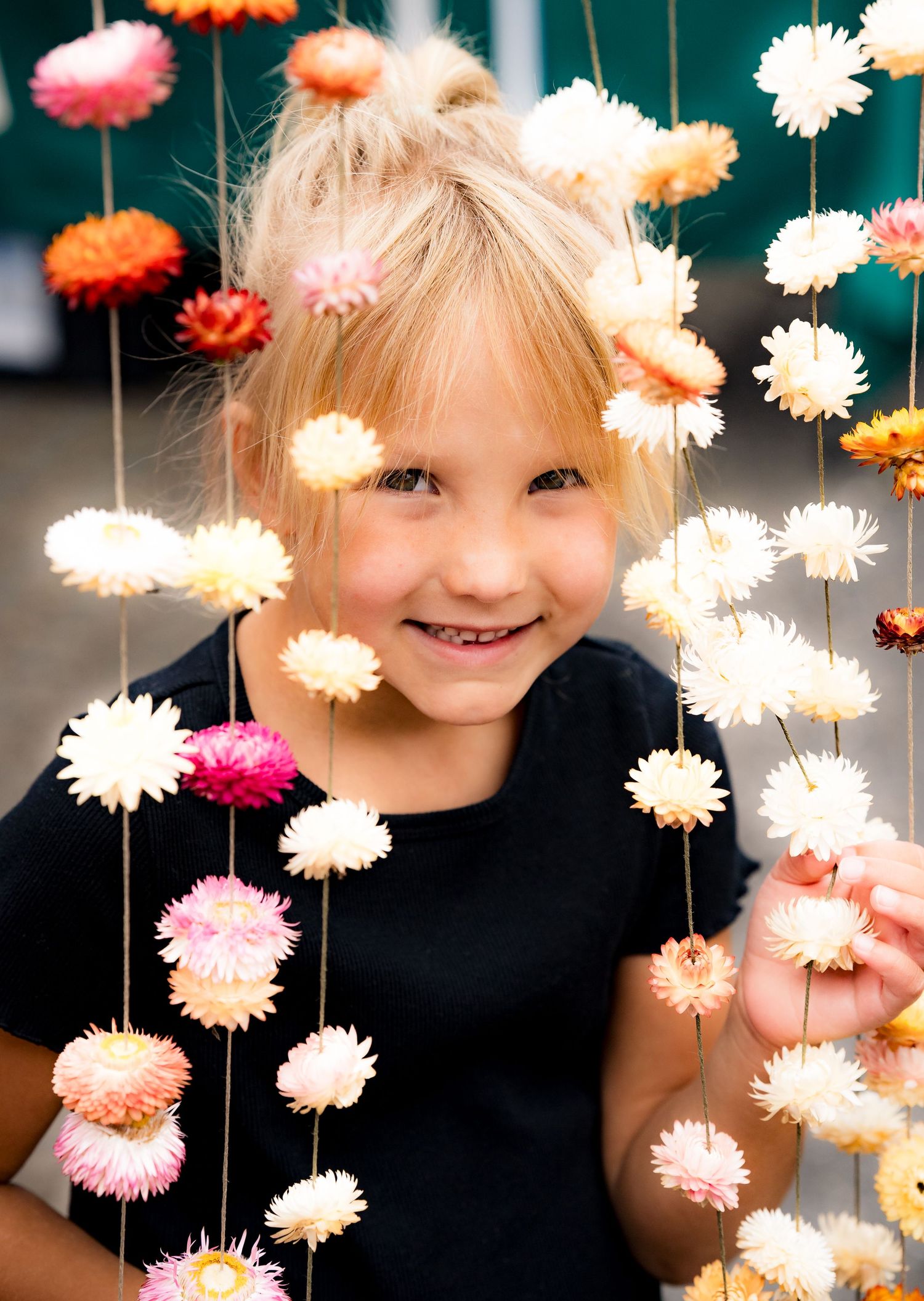 Smiling young blonde girl in black dress posing behind hanging floral garlands with pink and peach strawflowers.