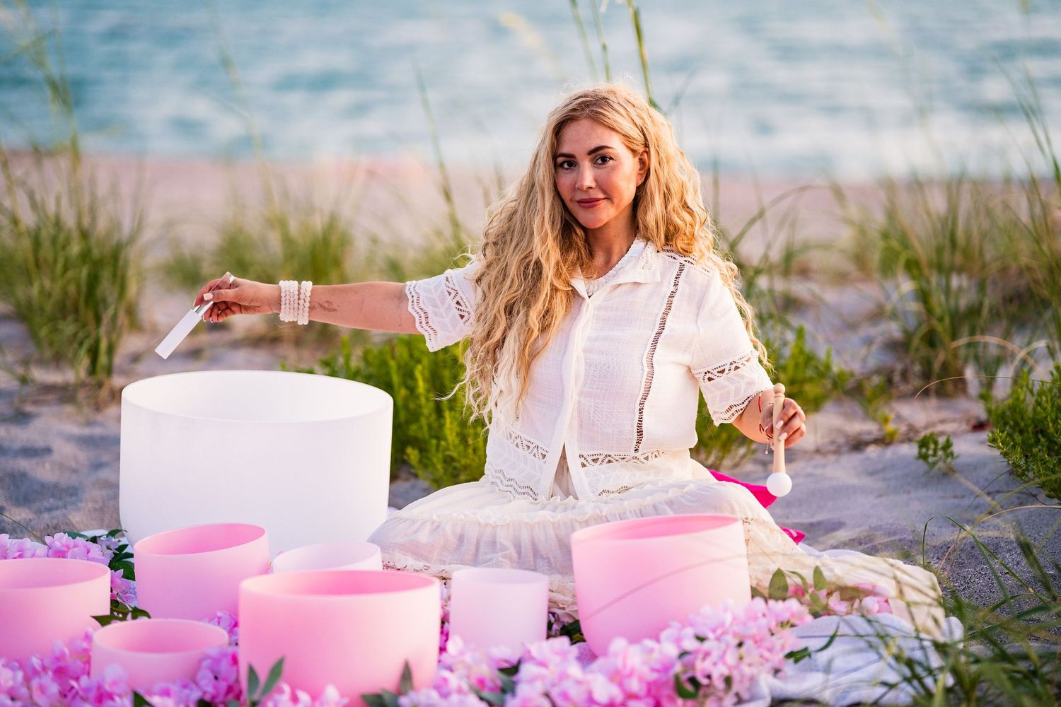 A photo of a blonde-haired woman playing the singing bowls, sitting peacefully on the beach.