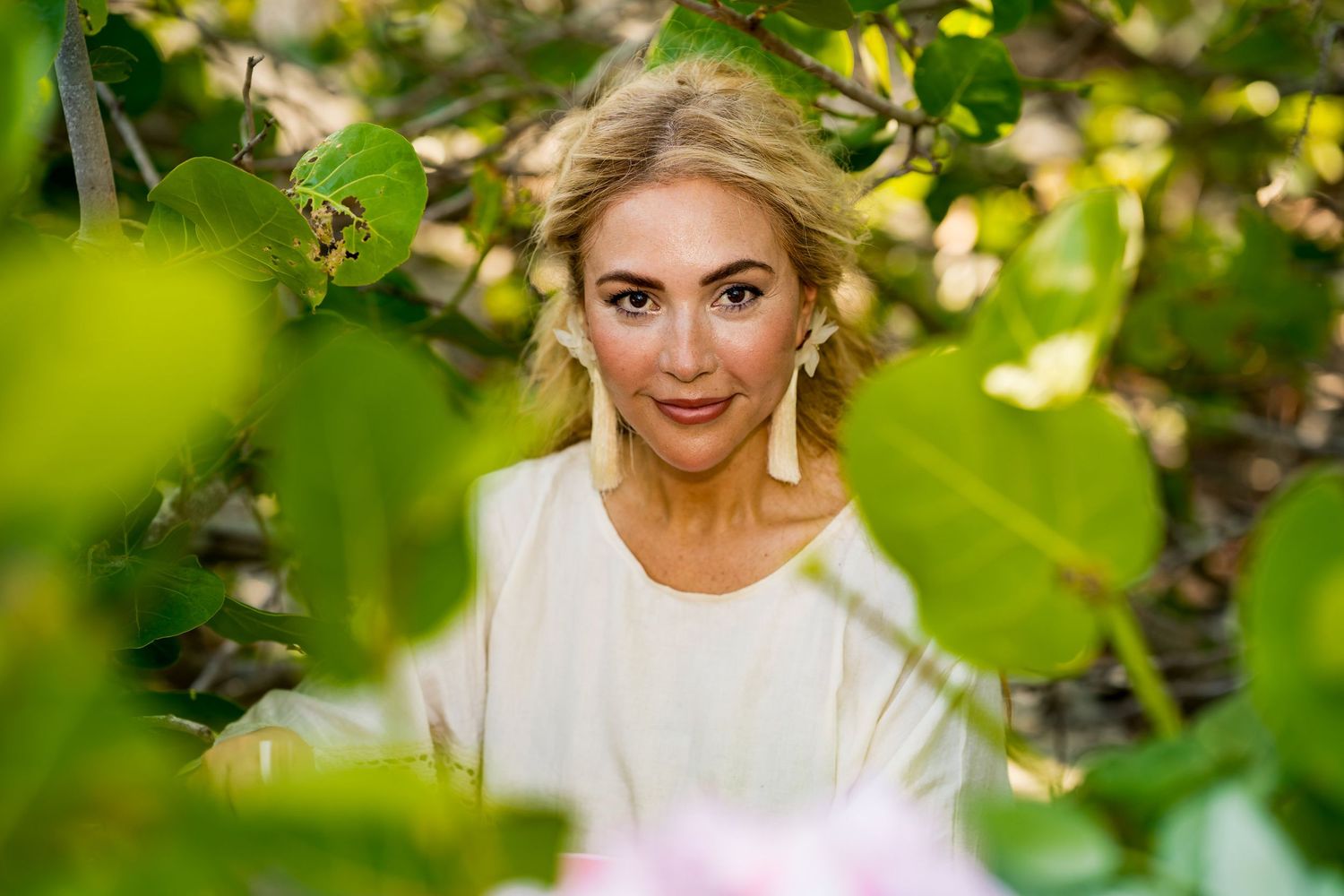 A photo of a blonde woman looking between green leaves, she is wearing white earrings and a white top.