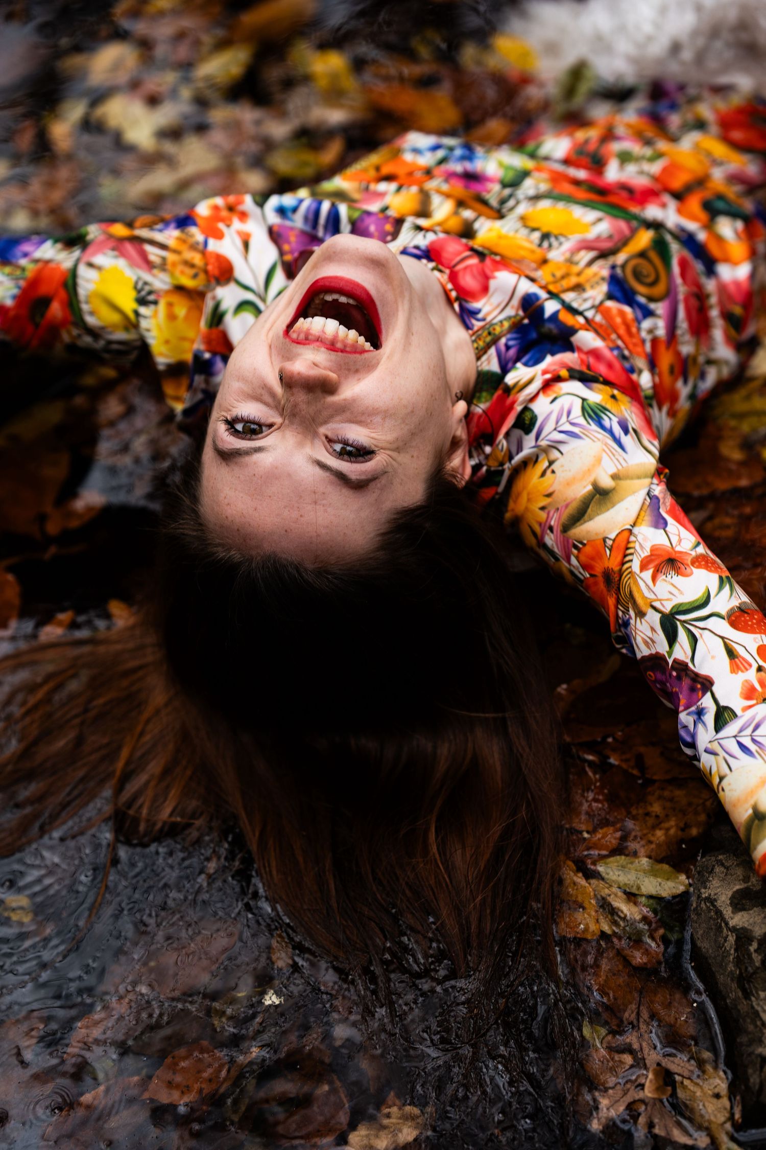 A person lying on autumn leaves wearing a colorful floral dress with their hair spread out against dark foliage.