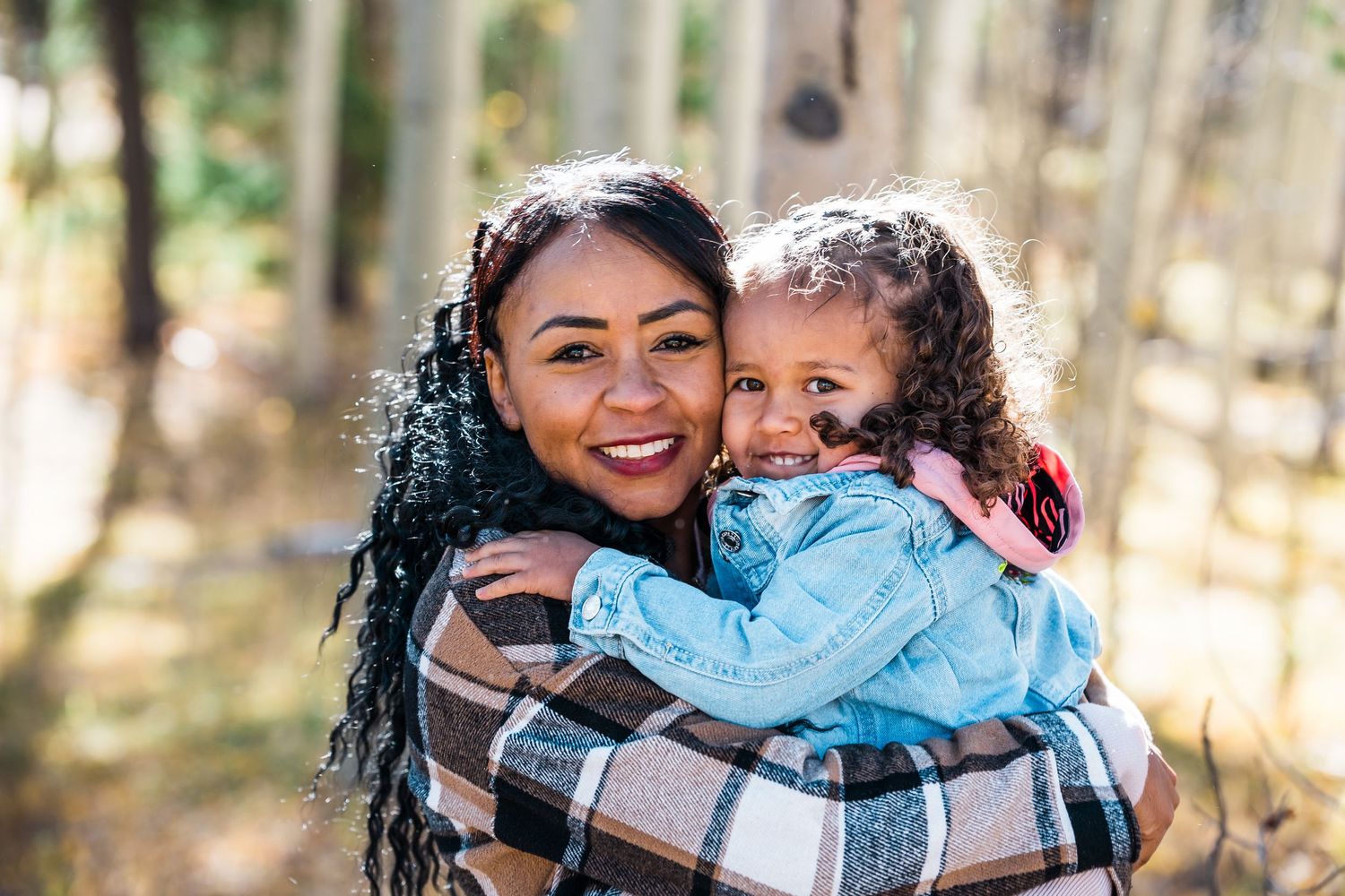 A photo of a woman in the woods with her little daughter hugging her and smiling at the camera.