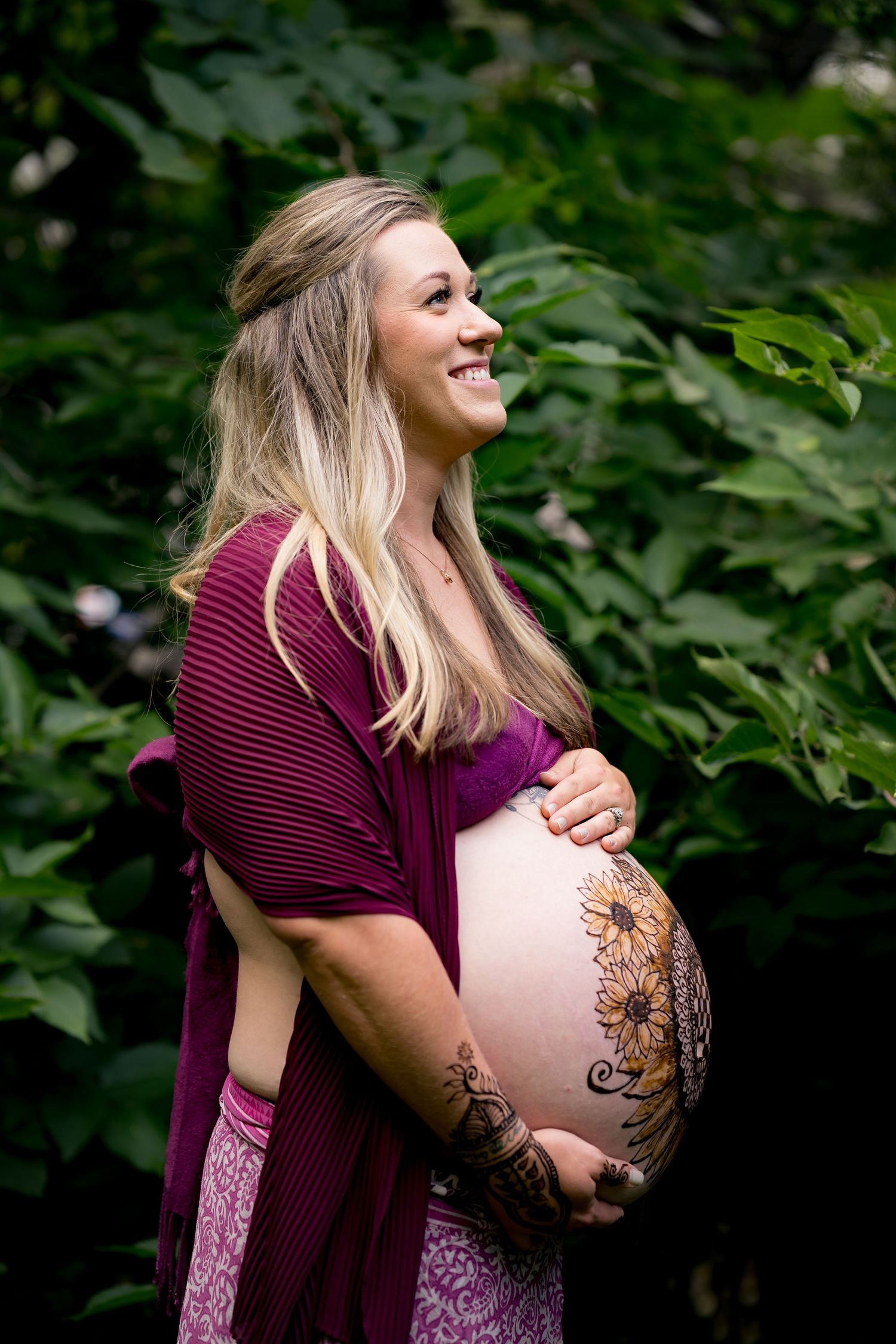 A pregnant woman in a burgundy dress laughs joyfully during a maternity photoshoot in a lush garden setting.