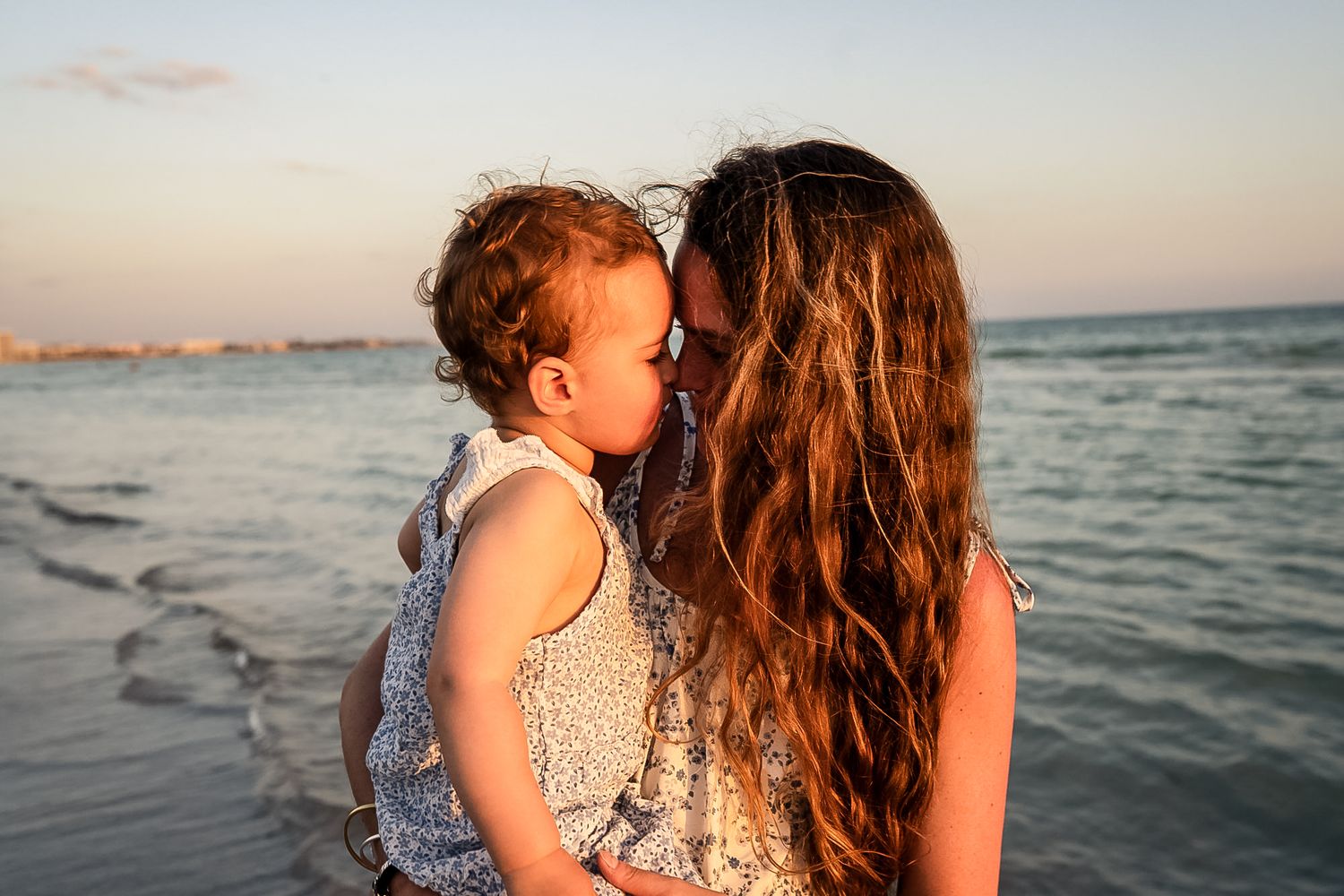 A photo of mother and daughter rubbing noses with the ocean behind them with beautiful sky behind them.