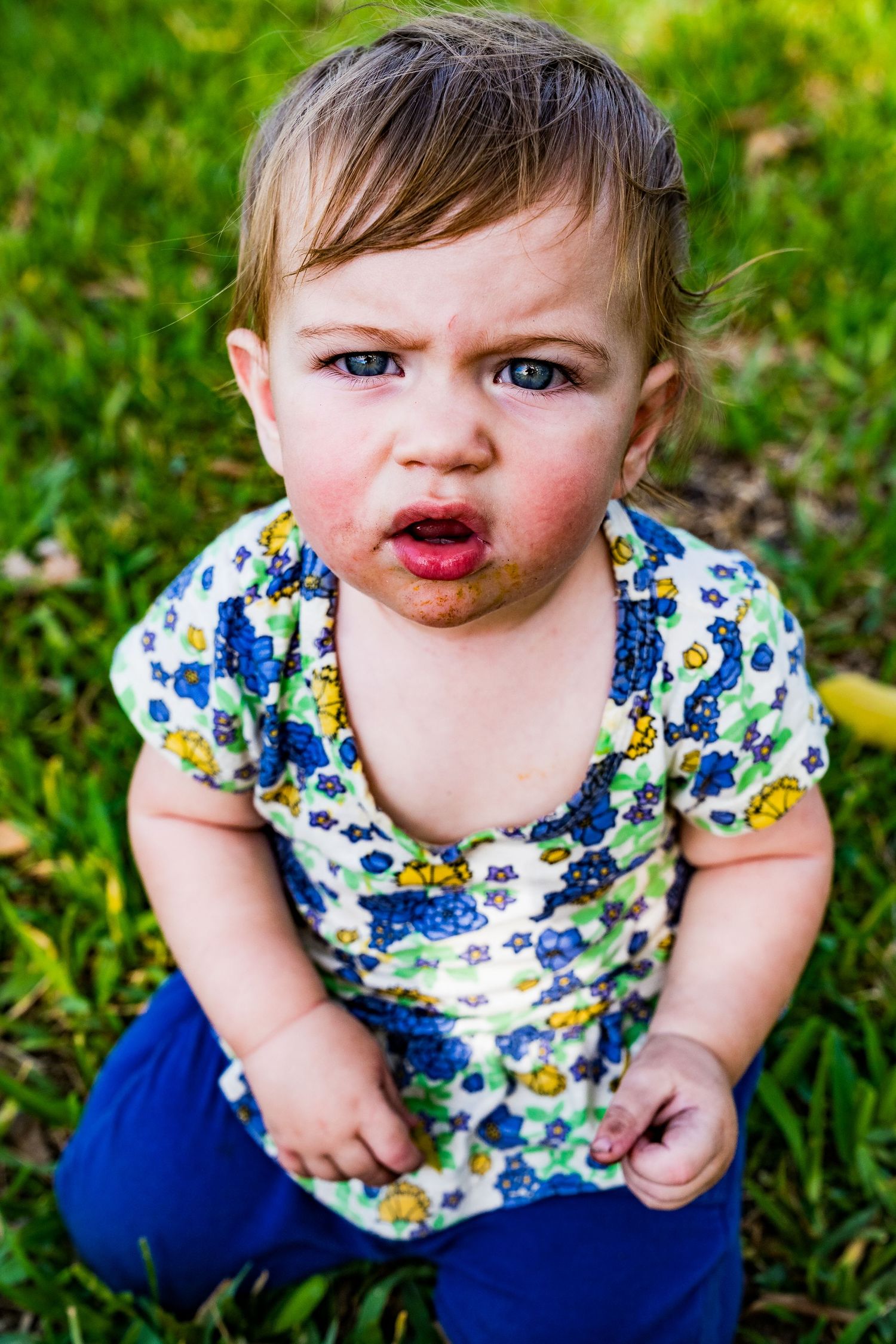 A photo of little girl with chunky arms by her sides looking intensely at the camera, grass is behind her.