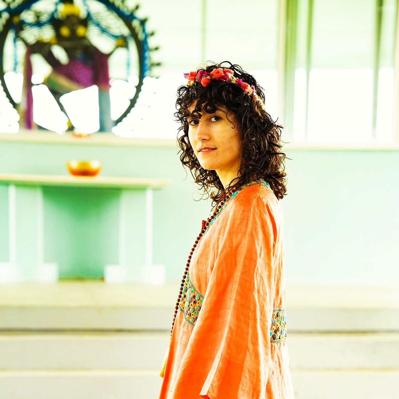 A photo of a woman wearing an orange outfit, wearing a flower crown with a deity statue behind her.