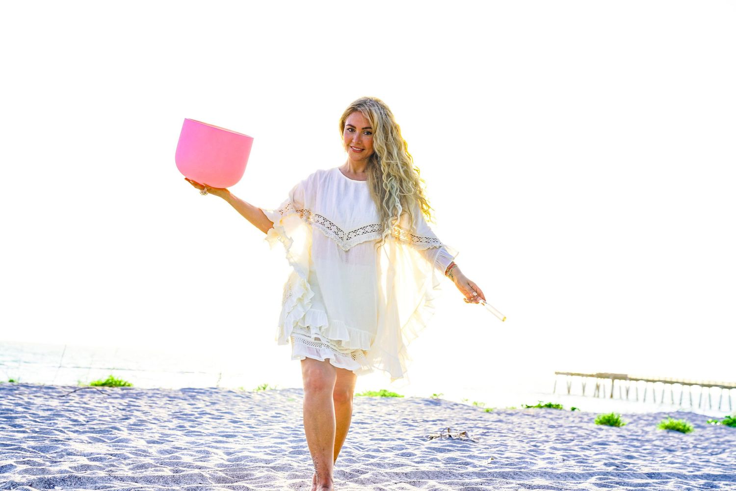 A photo of a woman with blonde wavy hair holding a pink bowl, she is walking on the sand on the beach.
