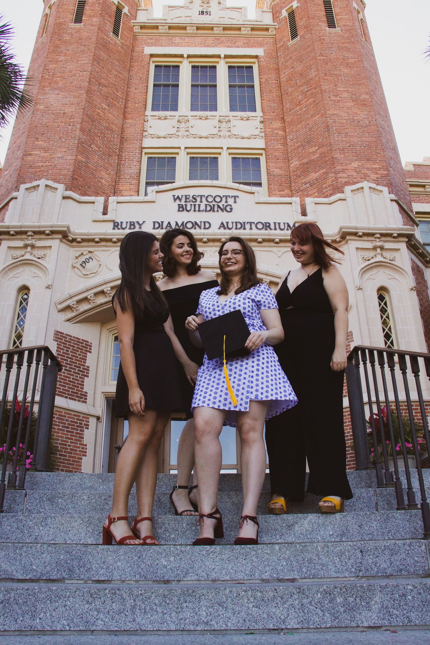 Four women pose on steps outside Westcott Building and Ruby Diamond Auditorium, one holding a graduation cap.