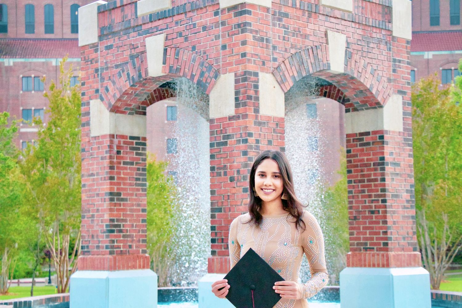 Smiling graduate holding cap in front of red brick arch fountain at university campus.