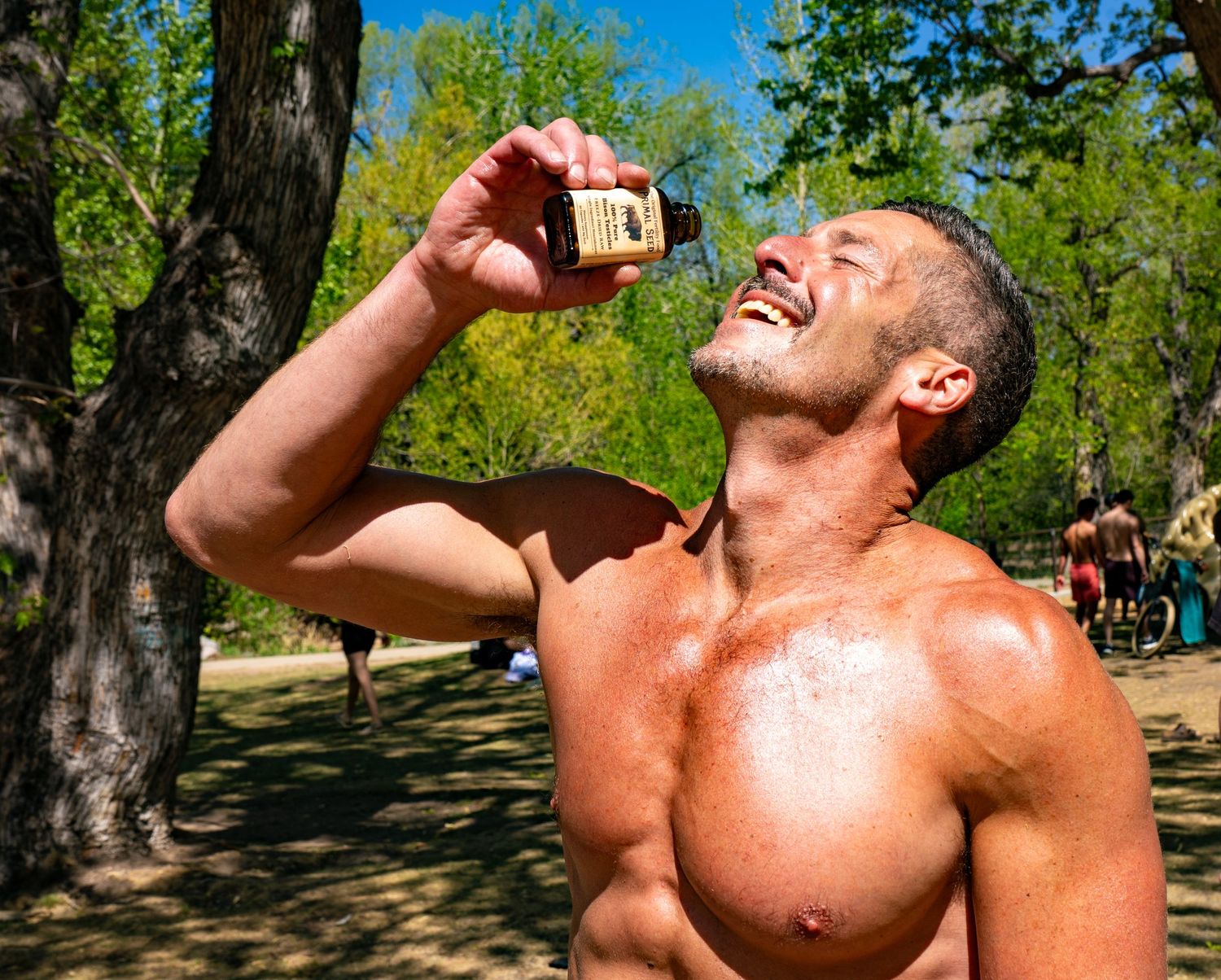 Muscular shirtless man laughing while drinking from a small bottle outdoors in a sunny park setting.