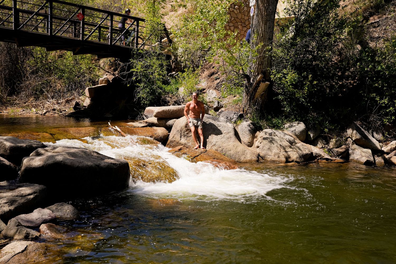 Person sitting on rocks beside a flowing stream with a wooden bridge and lush trees in background.
