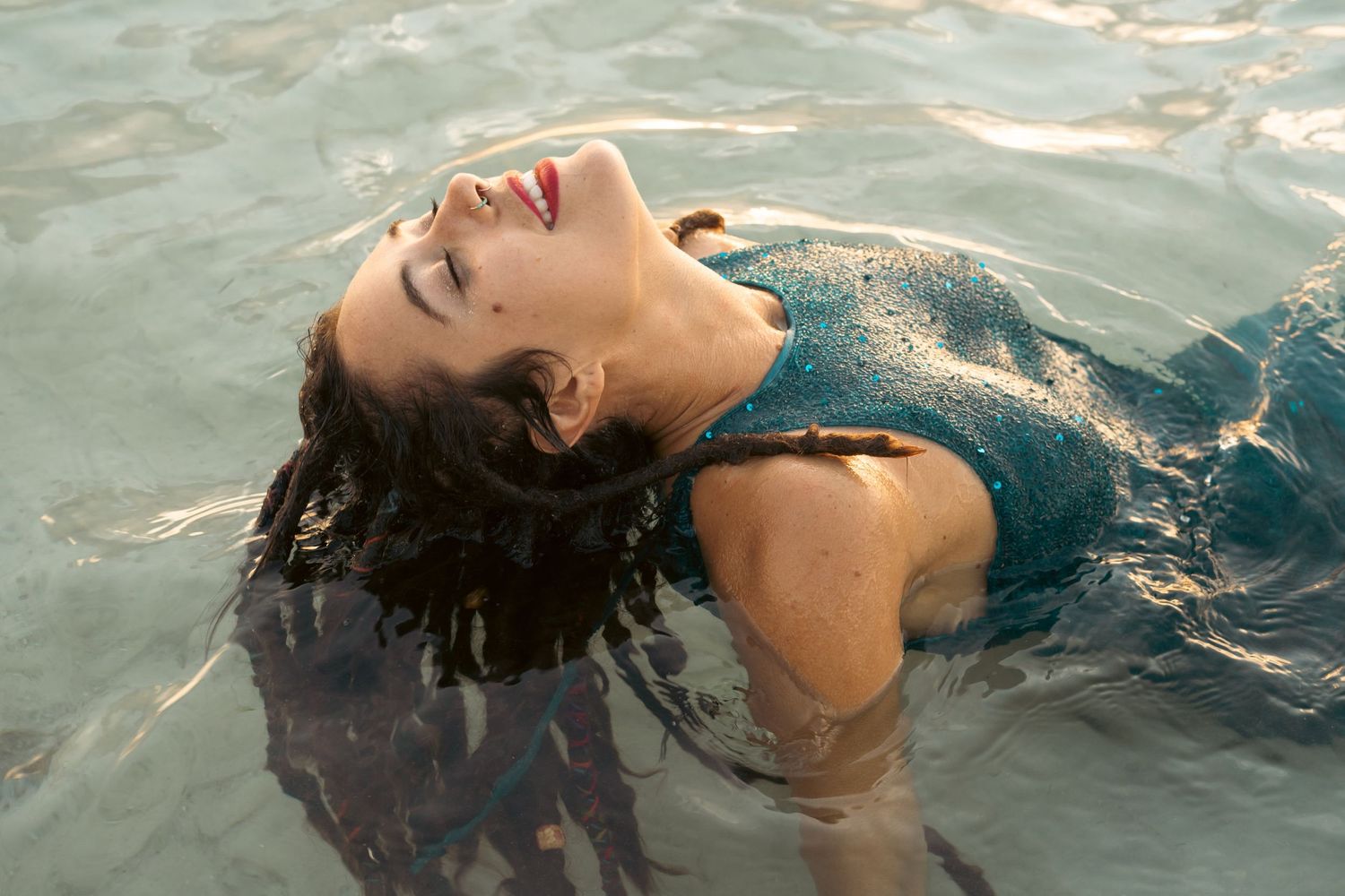 Woman in teal swimsuit floating in pool, head tilted back, eyes closed, smiling with red lips.