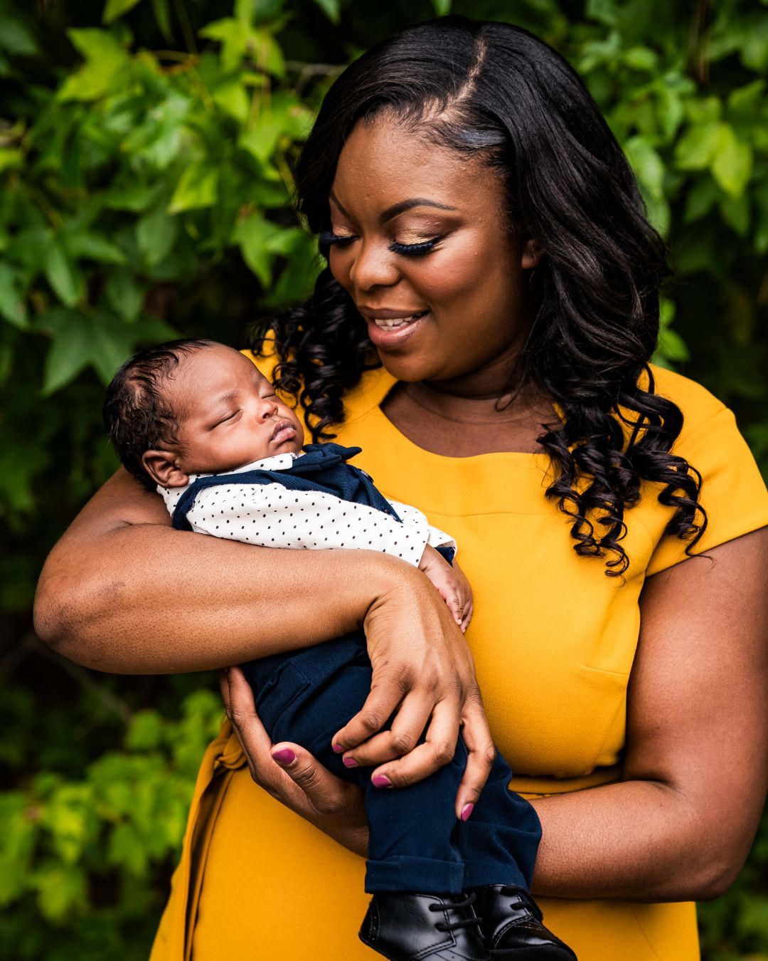 Smiling woman in yellow dress tenderly holds sleeping newborn baby outdoors with green leaves background.