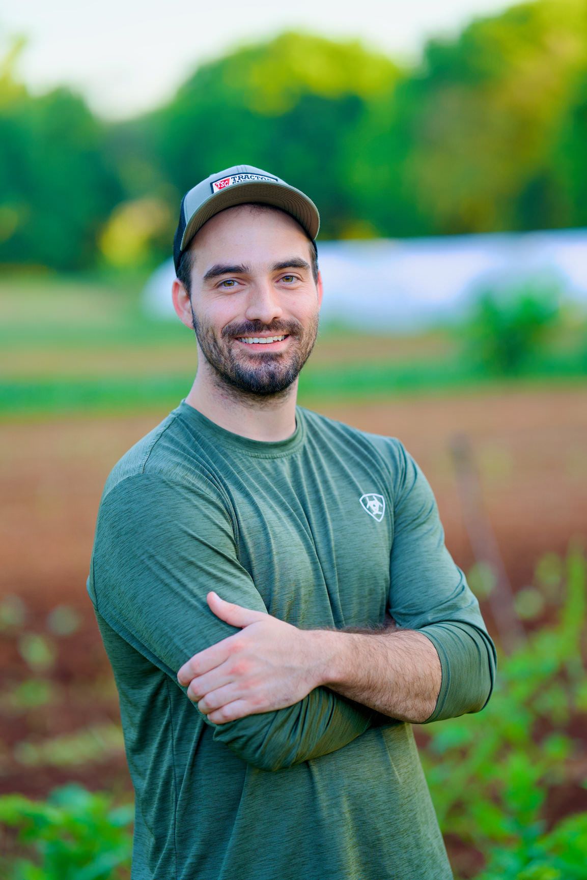 Smiling man with crossed arms standing in a green farm field wearing a cap and olive shirt.