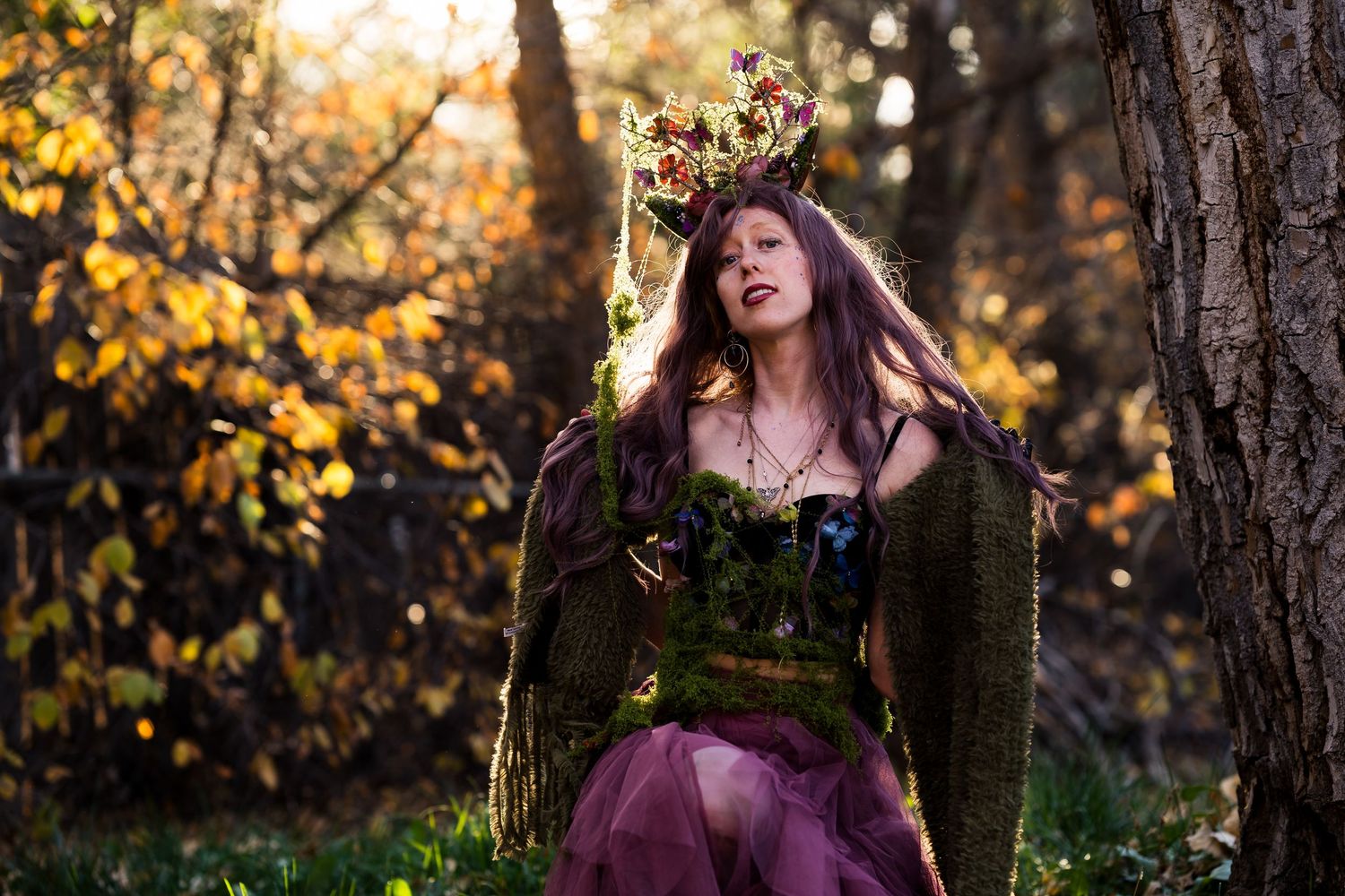 Woman in floral crown and forest fairy costume poses among autumn trees in golden sunlight.