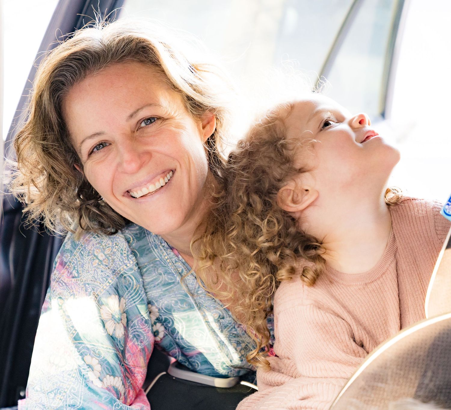 A parent and child with curly hair share a joyful moment while sitting together in natural light.