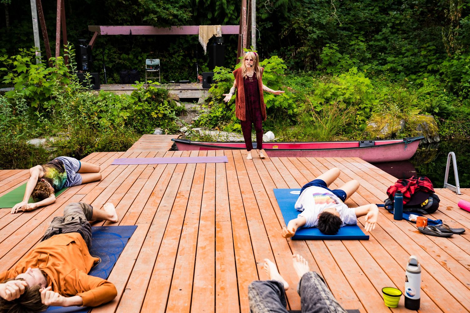 Yoga instructor leads an outdoor class on a wooden deck surrounded by lush forest, with students lying on colorful mats.
