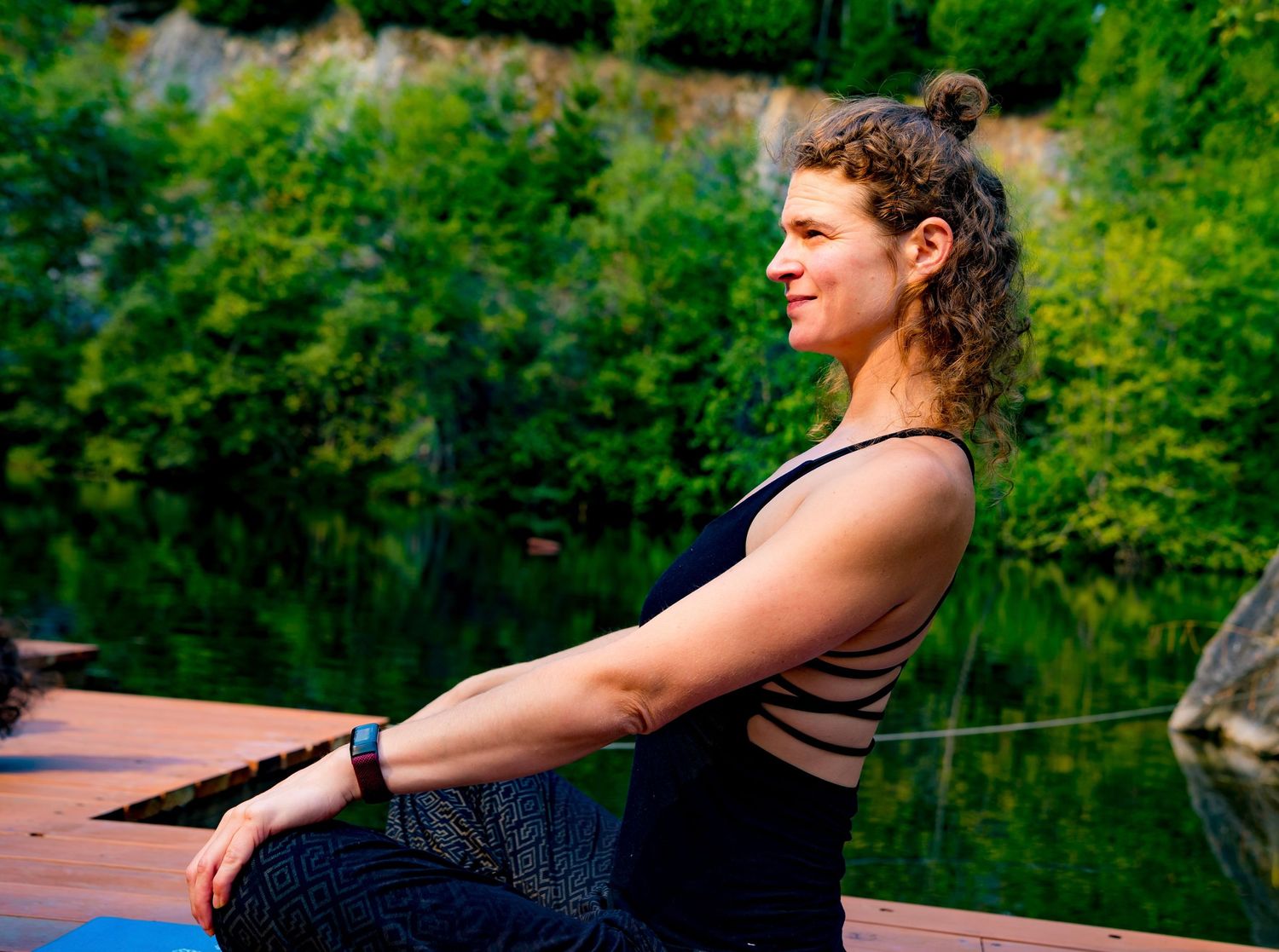 Woman practicing yoga meditation on a wooden dock by a serene green lake surrounded by lush trees.