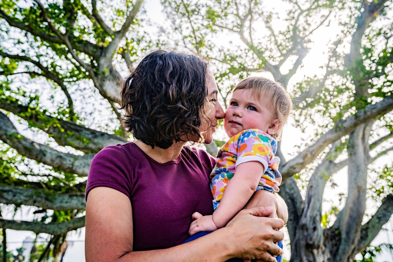A photo of a woman wearing a bright pink shirt, smiling at her baby girl with a tree behind them.