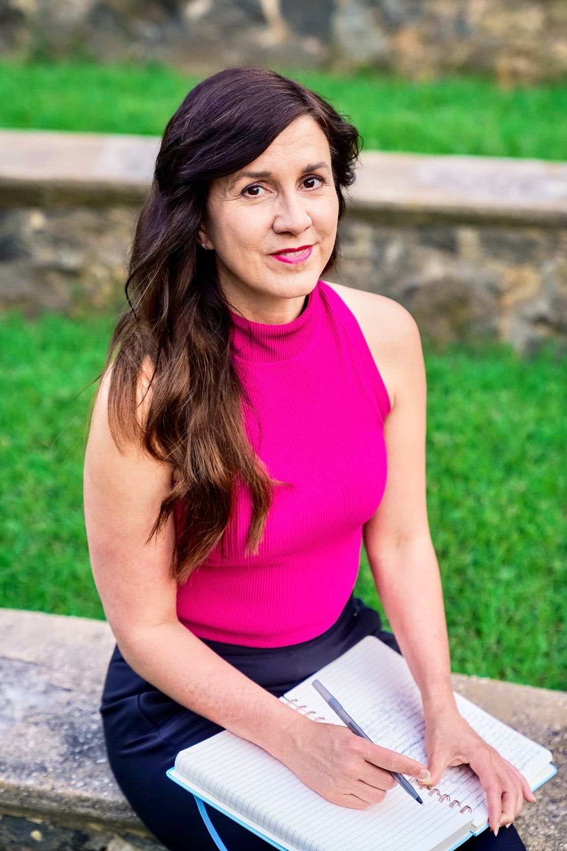 A photo of a woman wearing a hot pink shirt holding her notebook with greenery behind her.