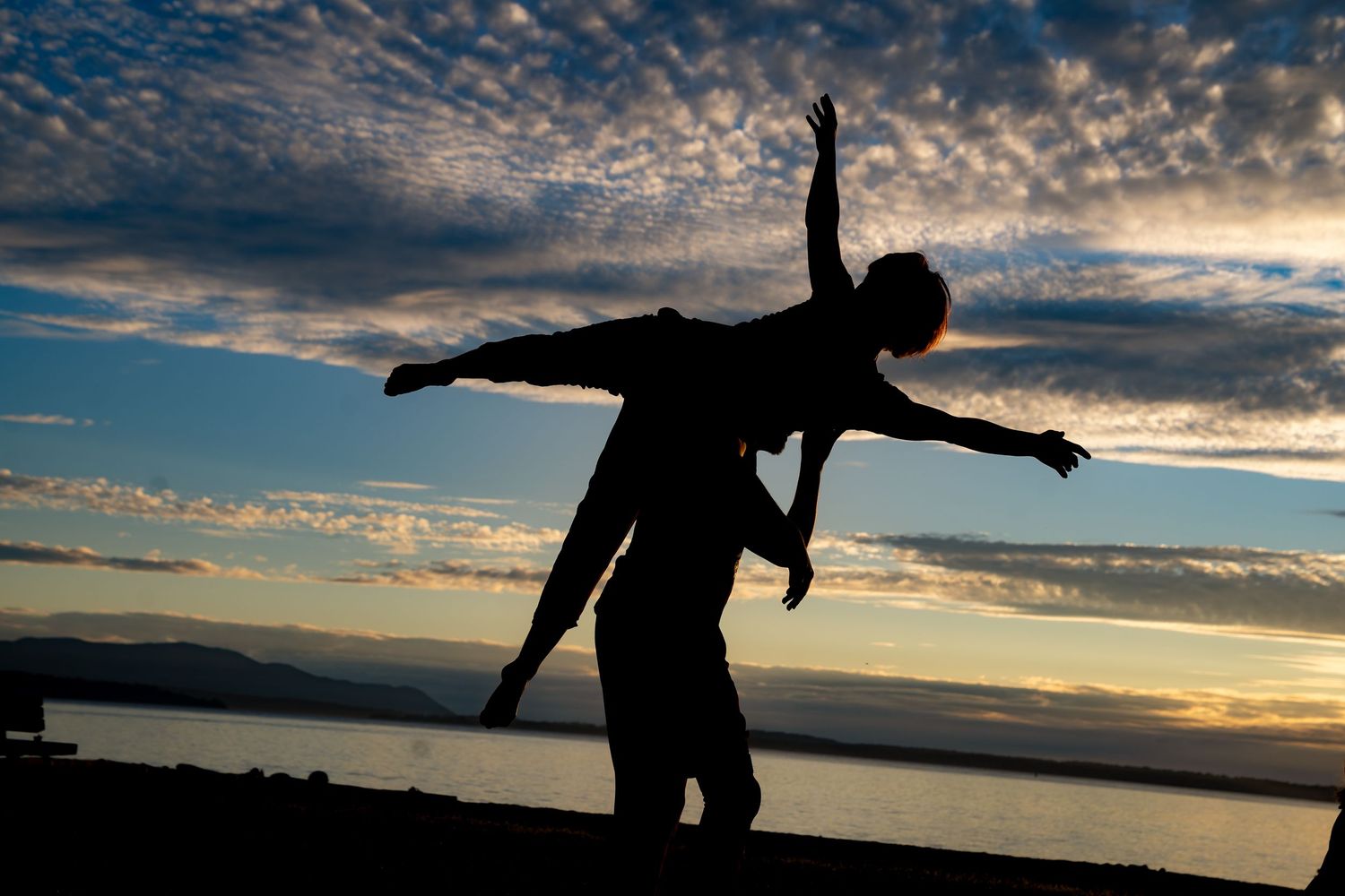 Silhouette of two dancers posing dramatically against a vibrant sunset sky with clouds over a calm ocean.