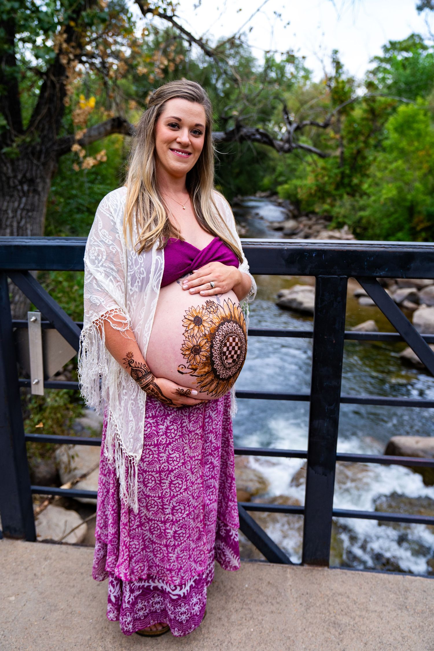 Woman in pink and purple dress with lace shawl poses near rushing waterfall and railing in outdoor setting.