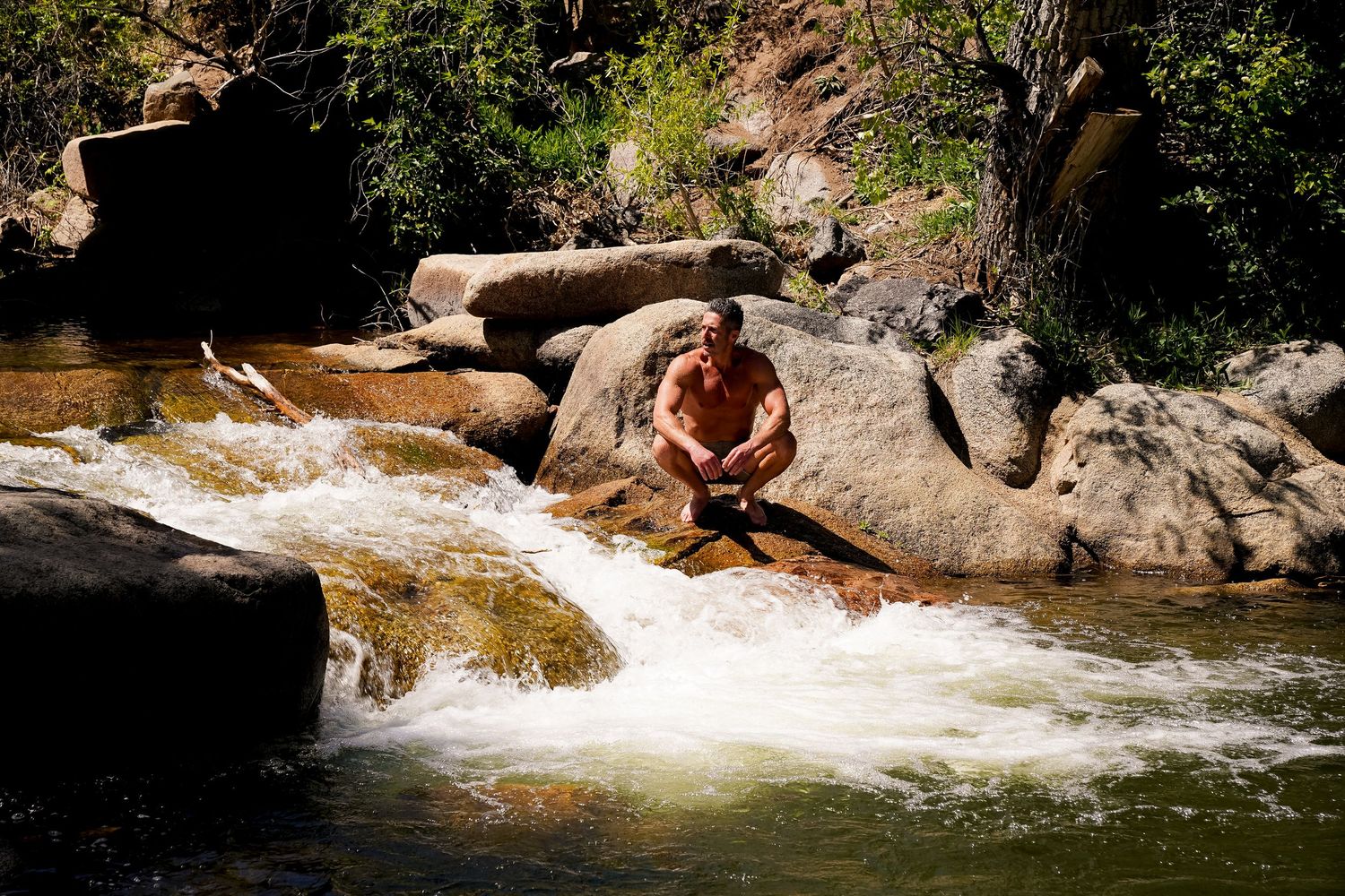 Man sitting on rocks beside a rushing mountain stream waterfall in a scenic natural wilderness setting.