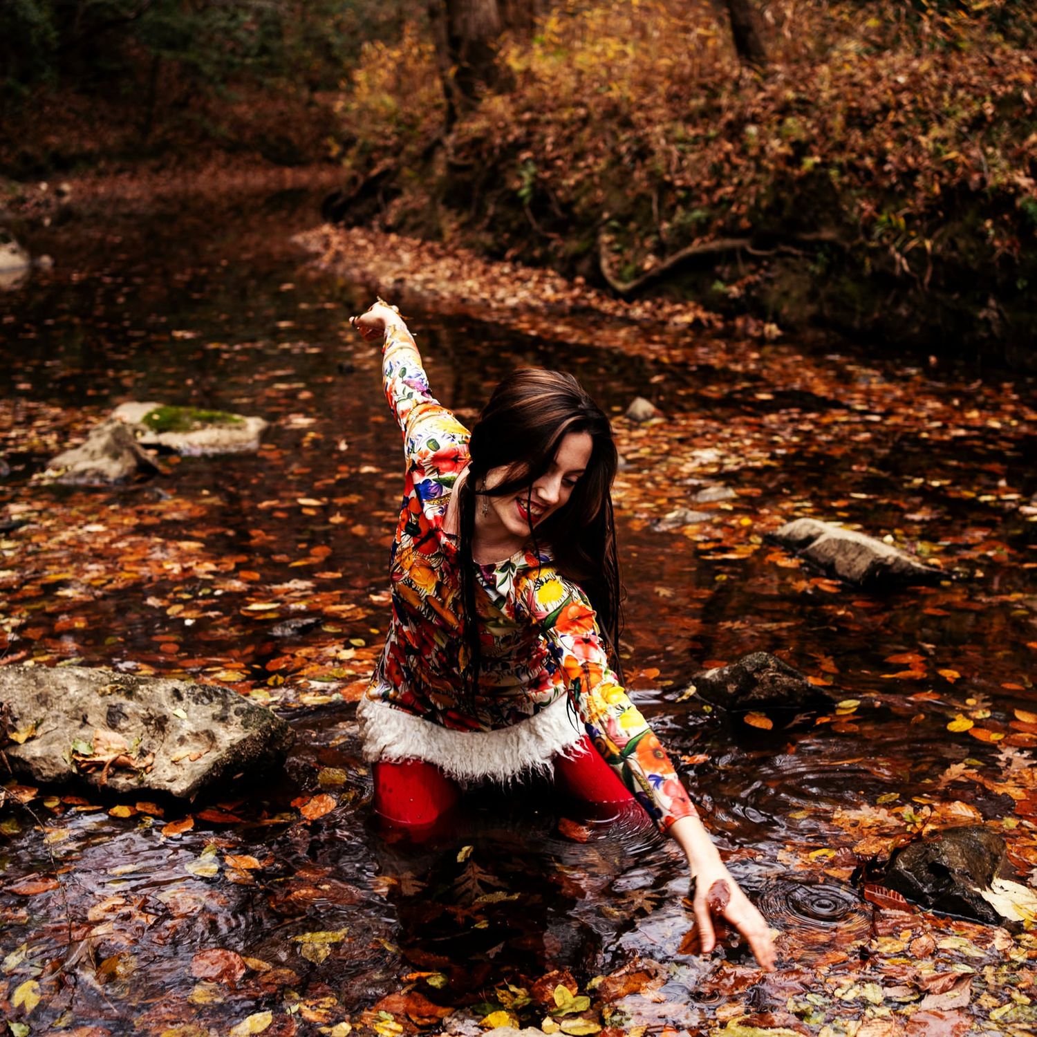 Woman in colorful floral outfit standing in autumn forest stream surrounded by fallen leaves.