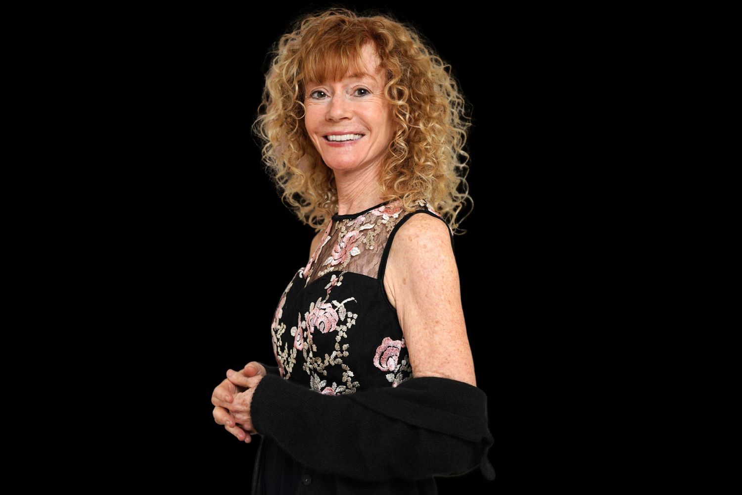A photo of a woman with red curly hair smiling at the camera, she is wear a black dress with flowers on it and is against a black backdrop.