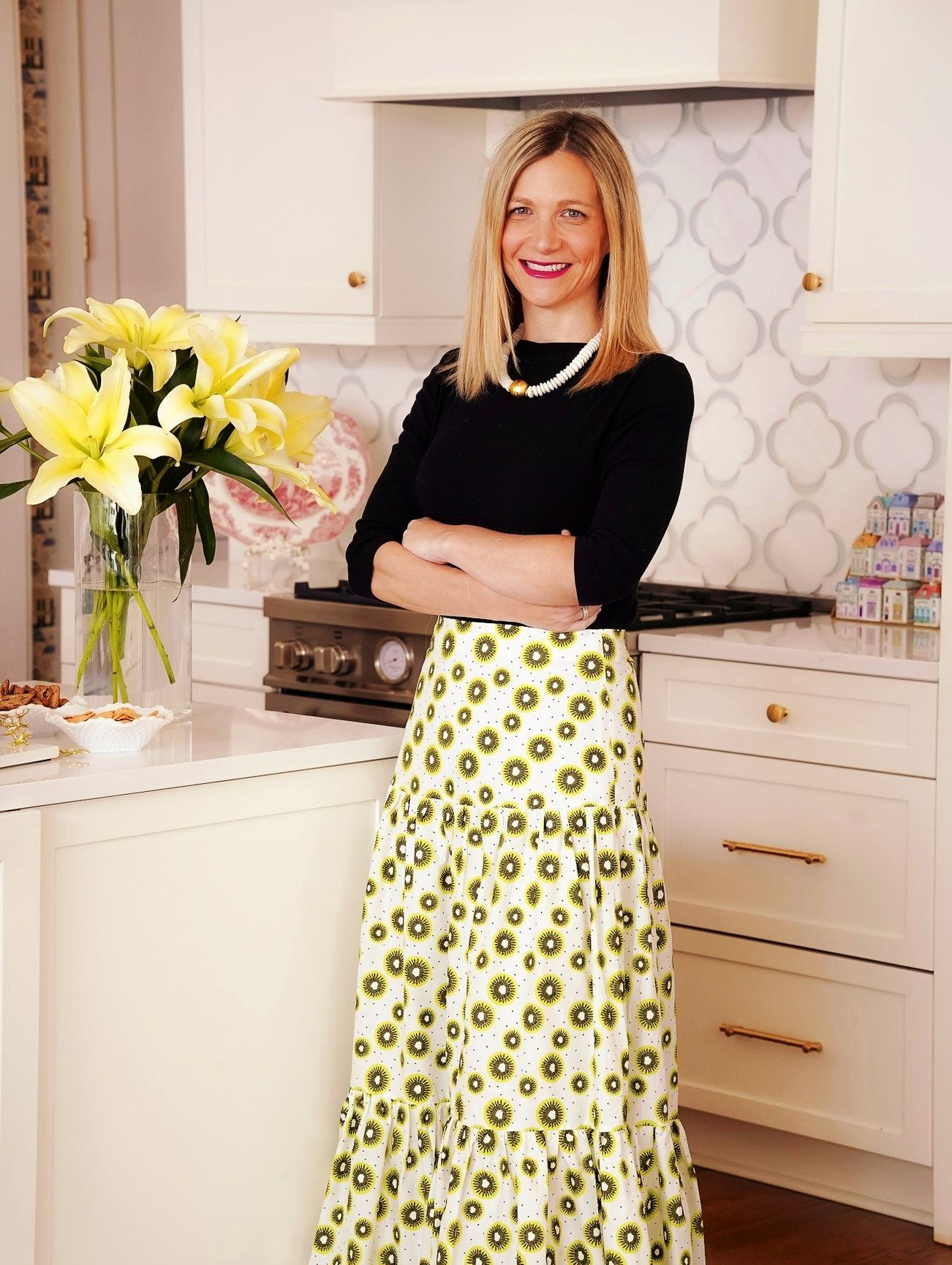 A photo of a woman wearing a black top, pearls and a long skirt with green polka dots and yellow flowers behind her.
